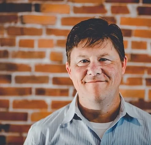 A man with short dark hair smiling in front of a brick wall.