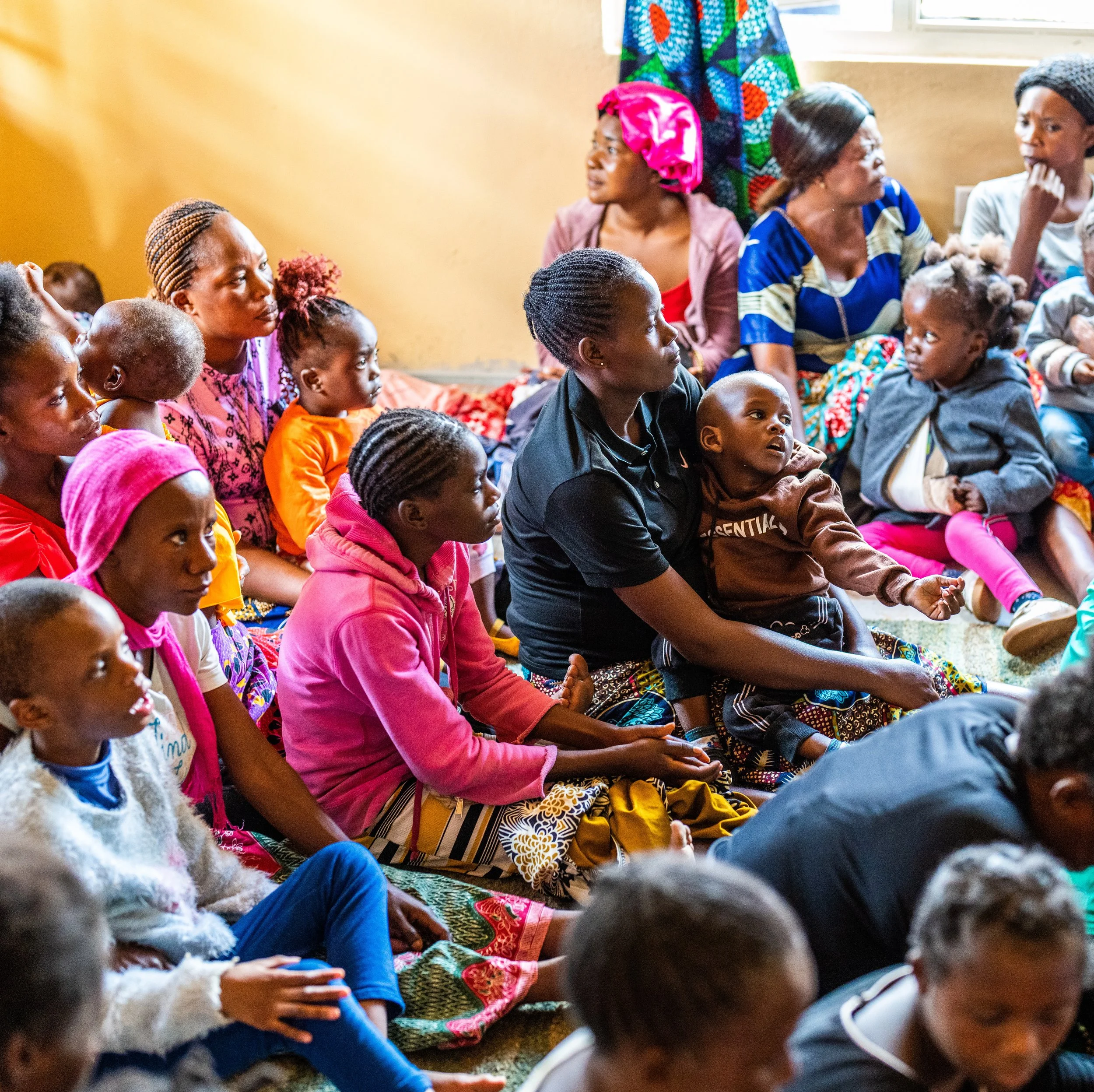 Group of women and children sitting on the floor in a room, attentively listening or watching something.