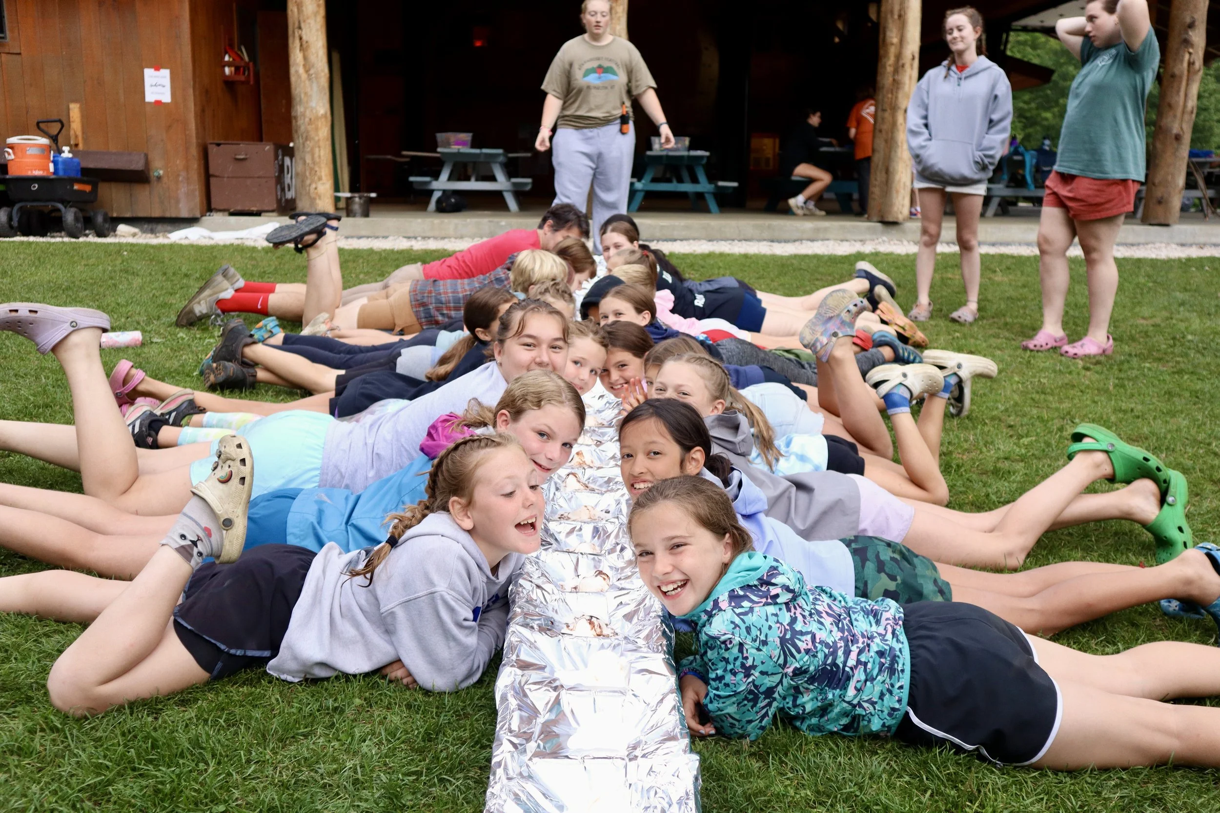 Group of children lying on grass during outdoor camp activity, smiling, some wearing casual clothes, with a metallic foil object in the center, and adults standing and sitting in the background.