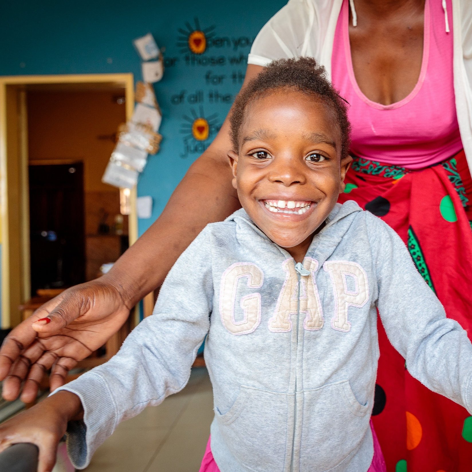 A young girl with a big smile wearing a gray hoodie, standing in front of a woman in a pink top and colorful skirt.
