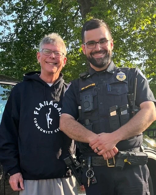 Two men, one in a hoodie and the other in police uniform, standing outdoors with trees in the background, smiling at the camera.