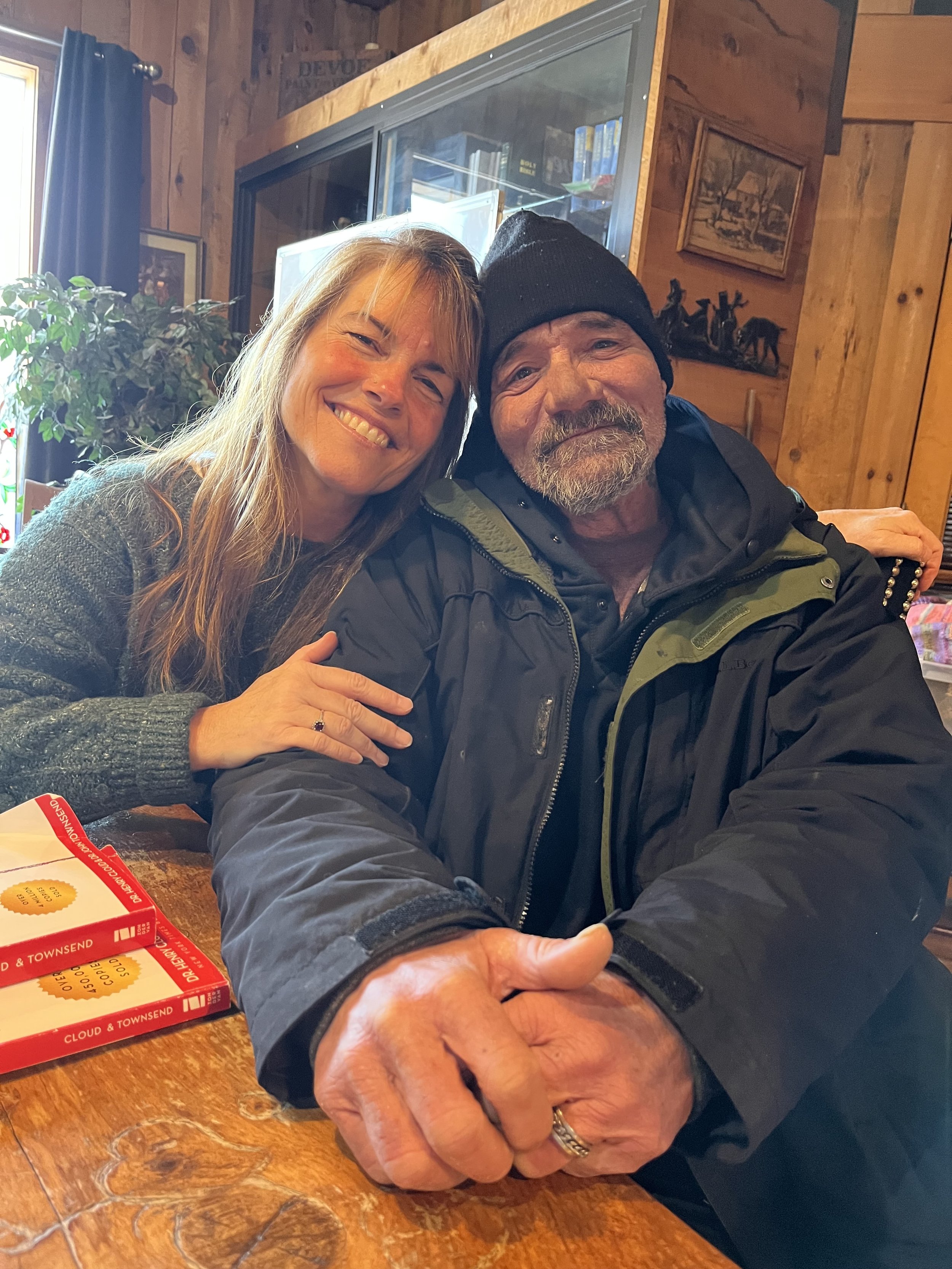 A smiling woman with light brown hair embracing a man wearing a black beanie and black jacket in a cozy wooden interior.