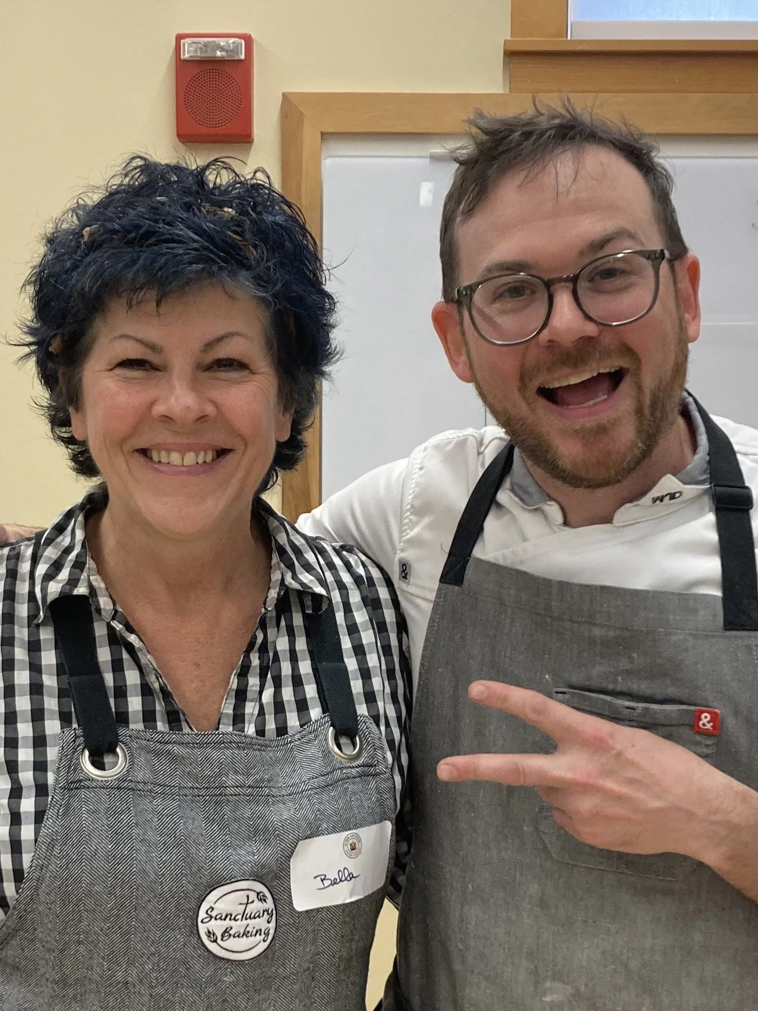 A woman and a man, both wearing aprons, smiling and standing close together indoors. The woman has short, curly dark hair and is wearing a checked shirt. The man has glasses, a beard, and is making a peace sign with his fingers.