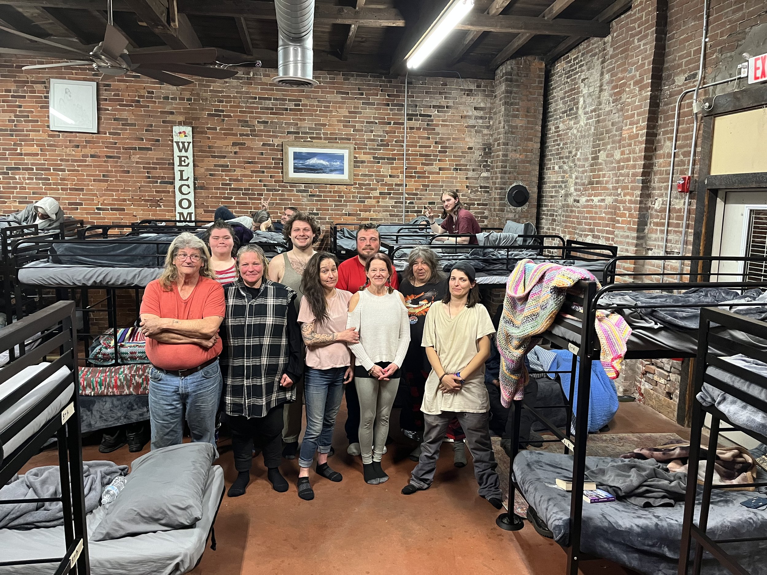 A group of ten people standing and smiling in a room with brick walls and bunk beds. There are several beds with blankets and personal items, and a 'Welcome' sign is visible on the wall.