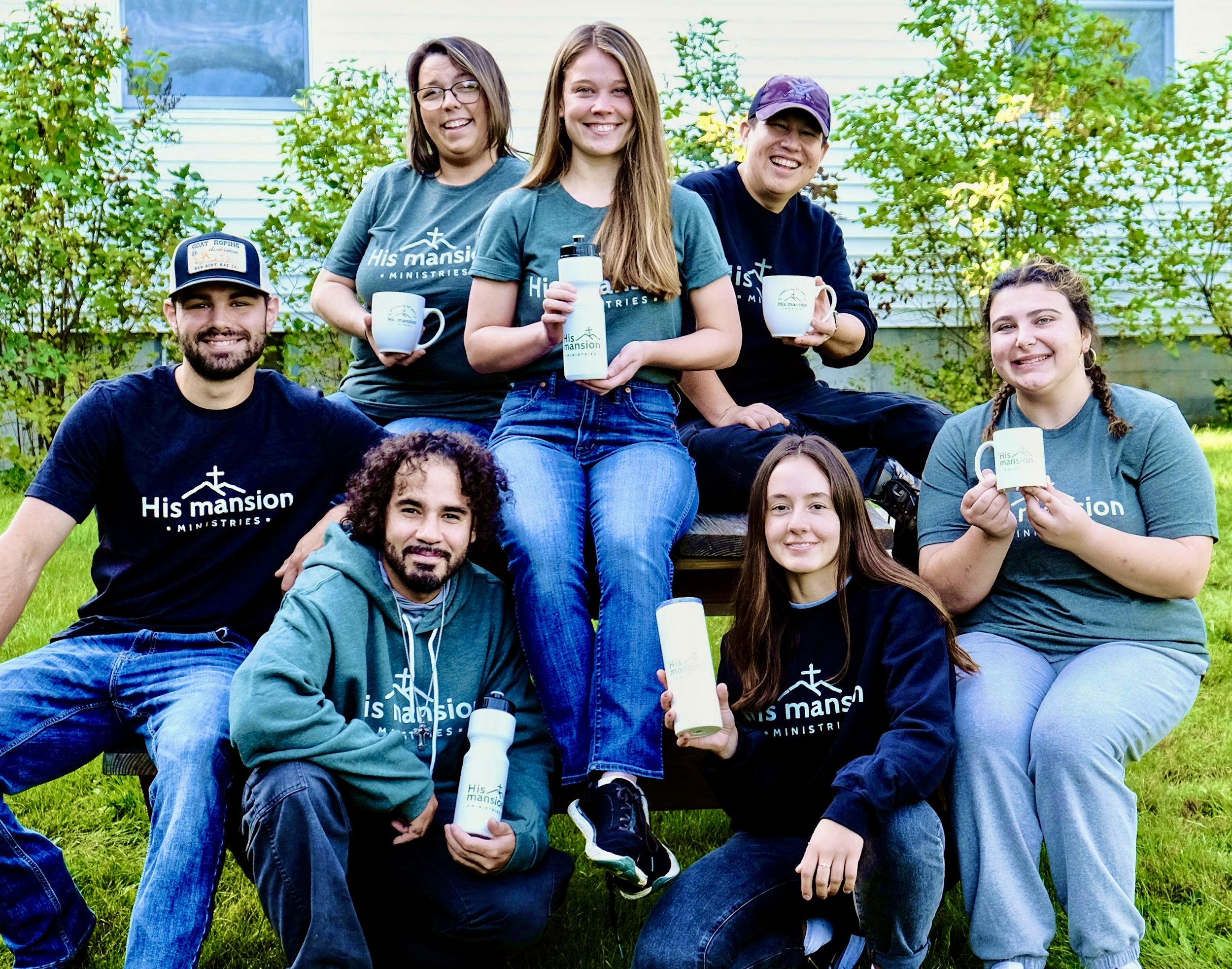 Group of eight young adults outdoors in a grassy yard, holding mugs and water bottles with 'His mansion' logo, smiling and sitting on and around a picnic table.