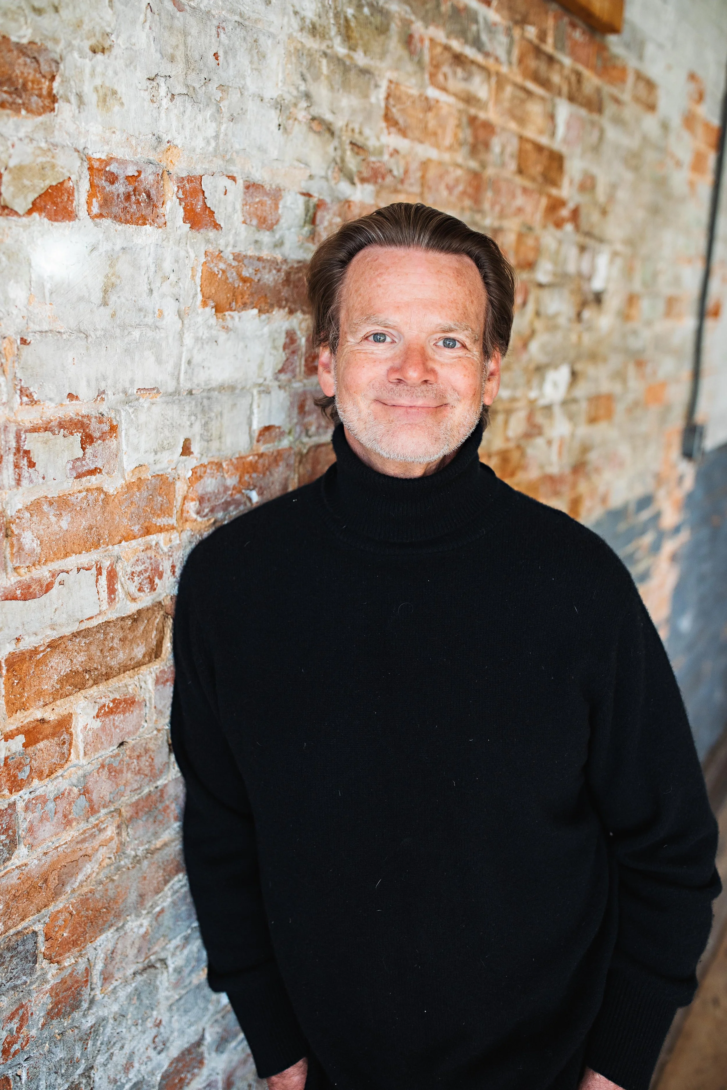 A middle-aged man with light skin, brown hair, and blue eyes smiling at the camera, wearing a black turtleneck sweater, standing against a rustic exposed brick wall with mixed red and white bricks.