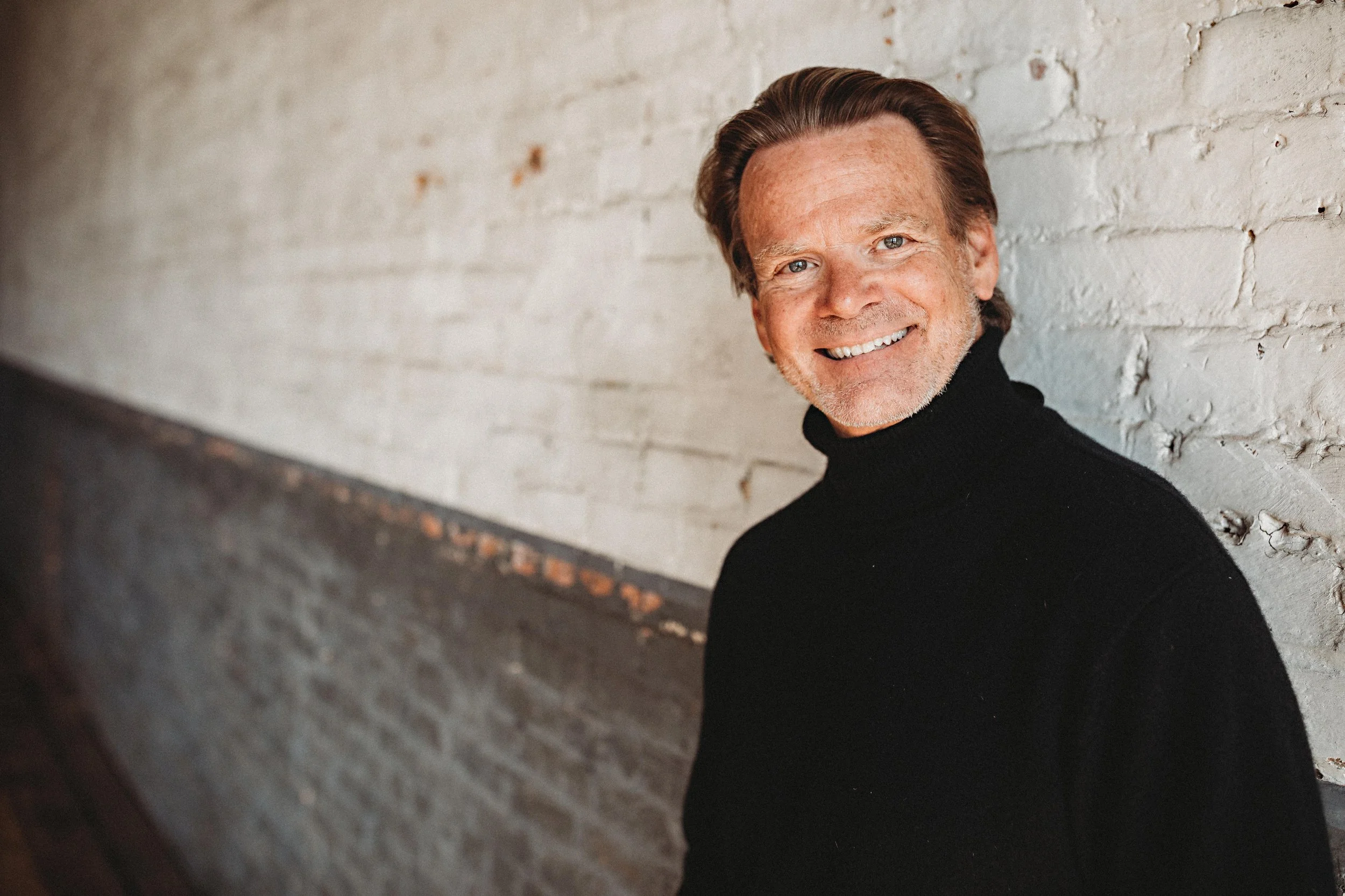 A close-up of a middle-aged man with gray hair, blue eyes, and a big smile, leaning against a white brick wall, wearing a black turtleneck sweater.