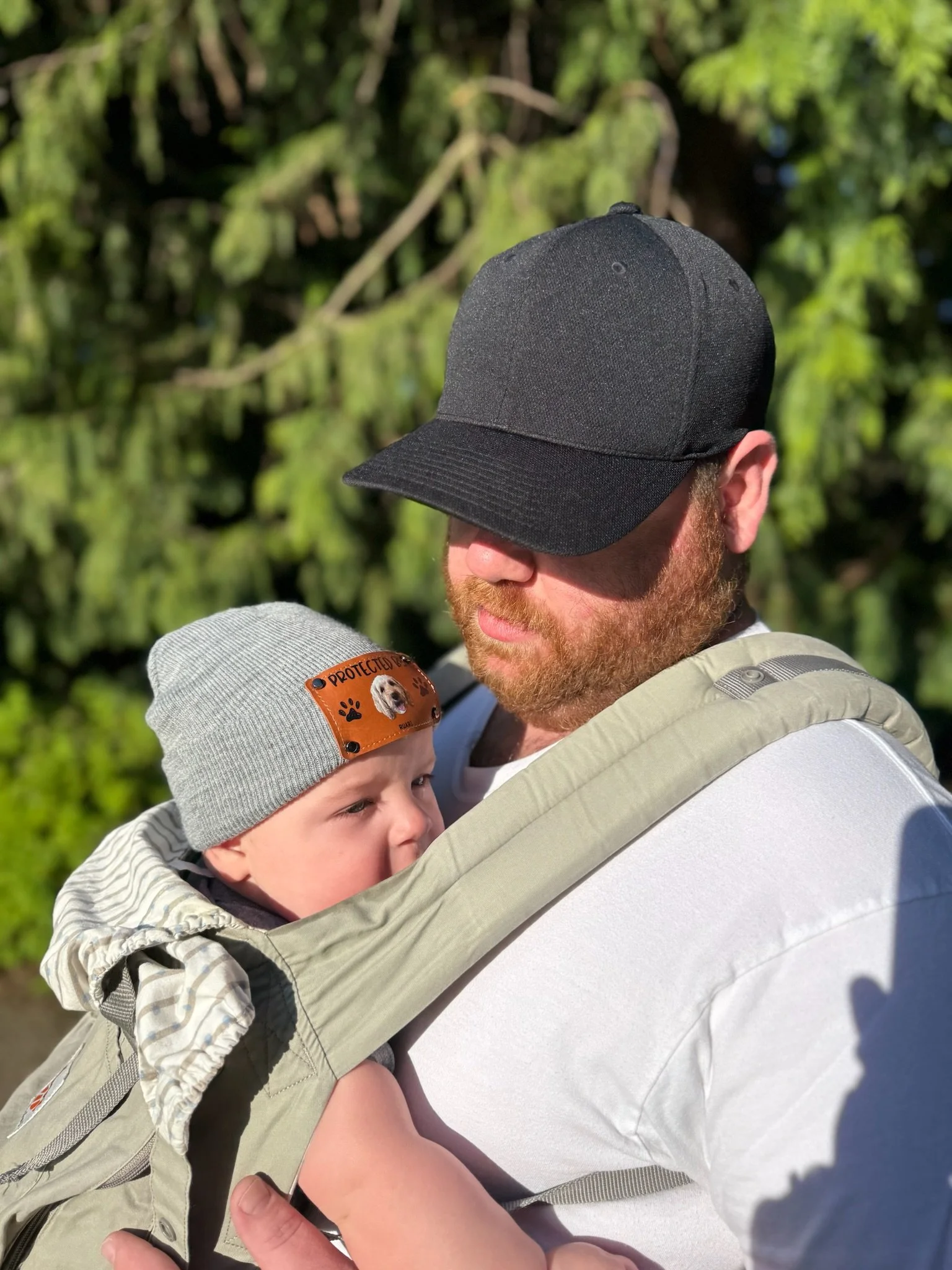 A man with a beard and a black cap holding a young child in a carrier outdoors, with a background of green trees.