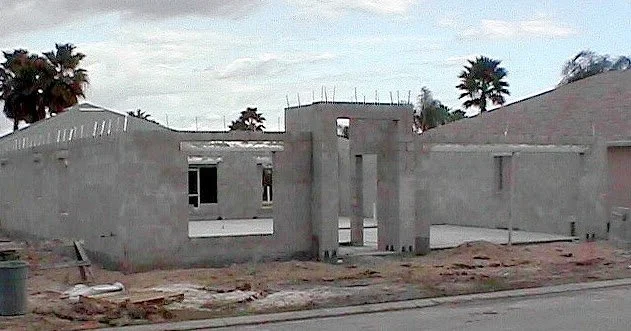 Under-construction concrete building structure on a dirt lot with palm trees and cloudy sky in the background.
