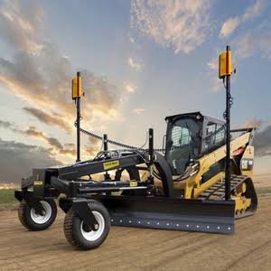 A large road construction vehicle with front blade, mounted on a dirt surface, under a partly cloudy sky at sunset.