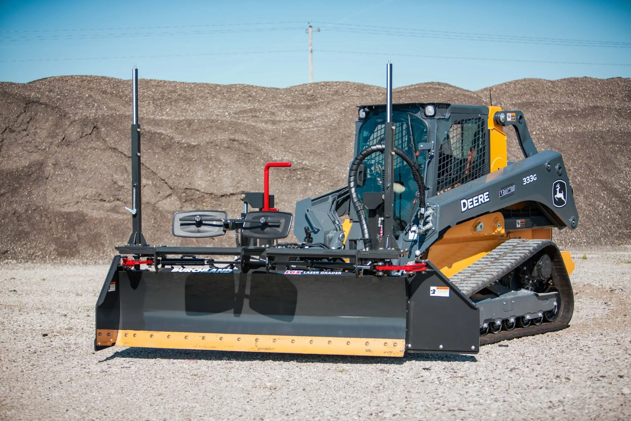 A compact track loader with a laser grading attachment working on a construction site with dirt mounds and electrical poles in the background.