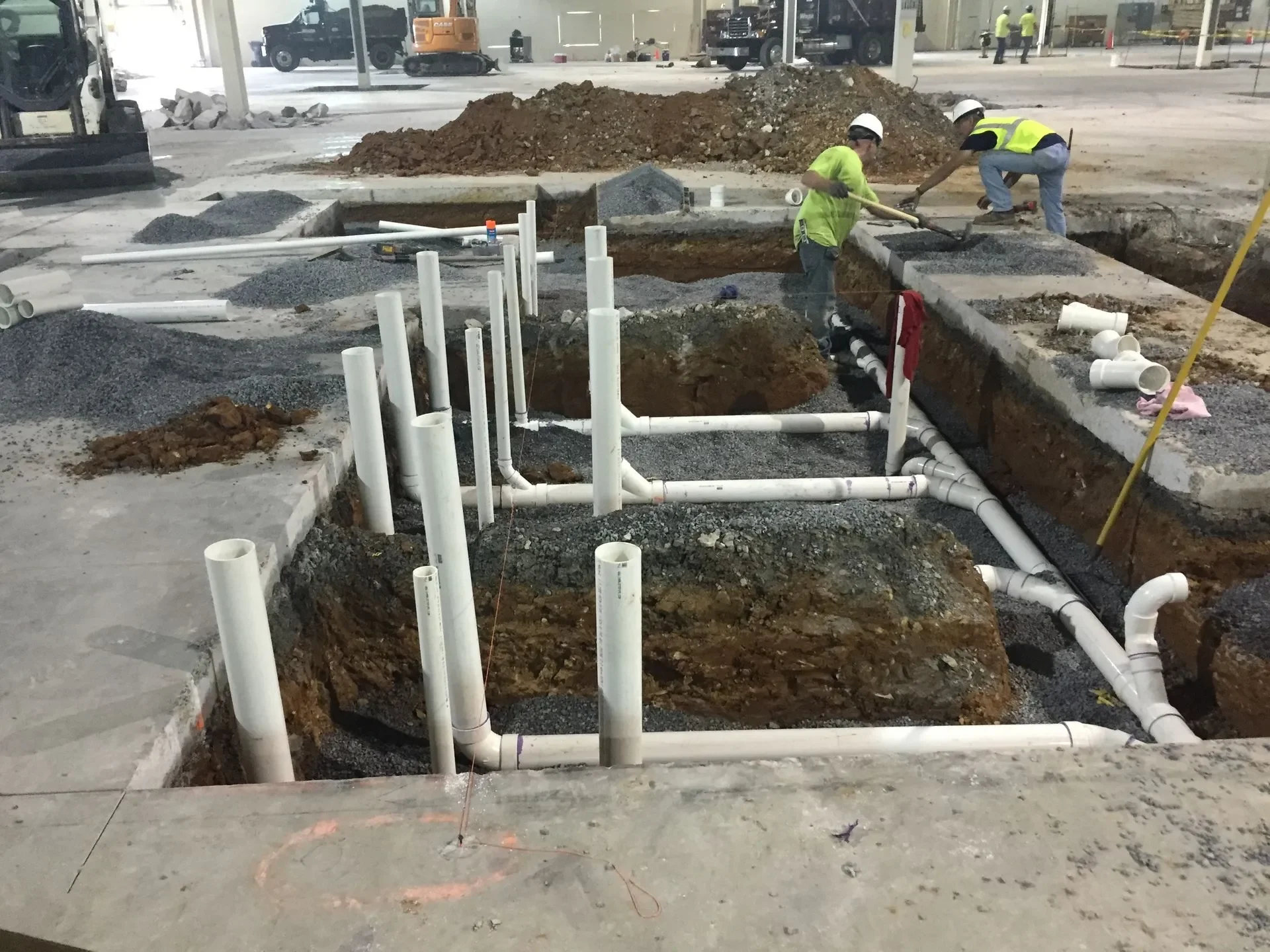Construction workers installing plumbing pipes in excavated trenches inside a building under construction, with construction equipment and vehicles in the background.