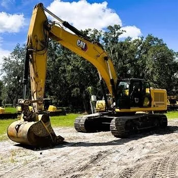 A yellow Caterpillar excavator parked on a dirt surface with trees and a blue sky in the background.