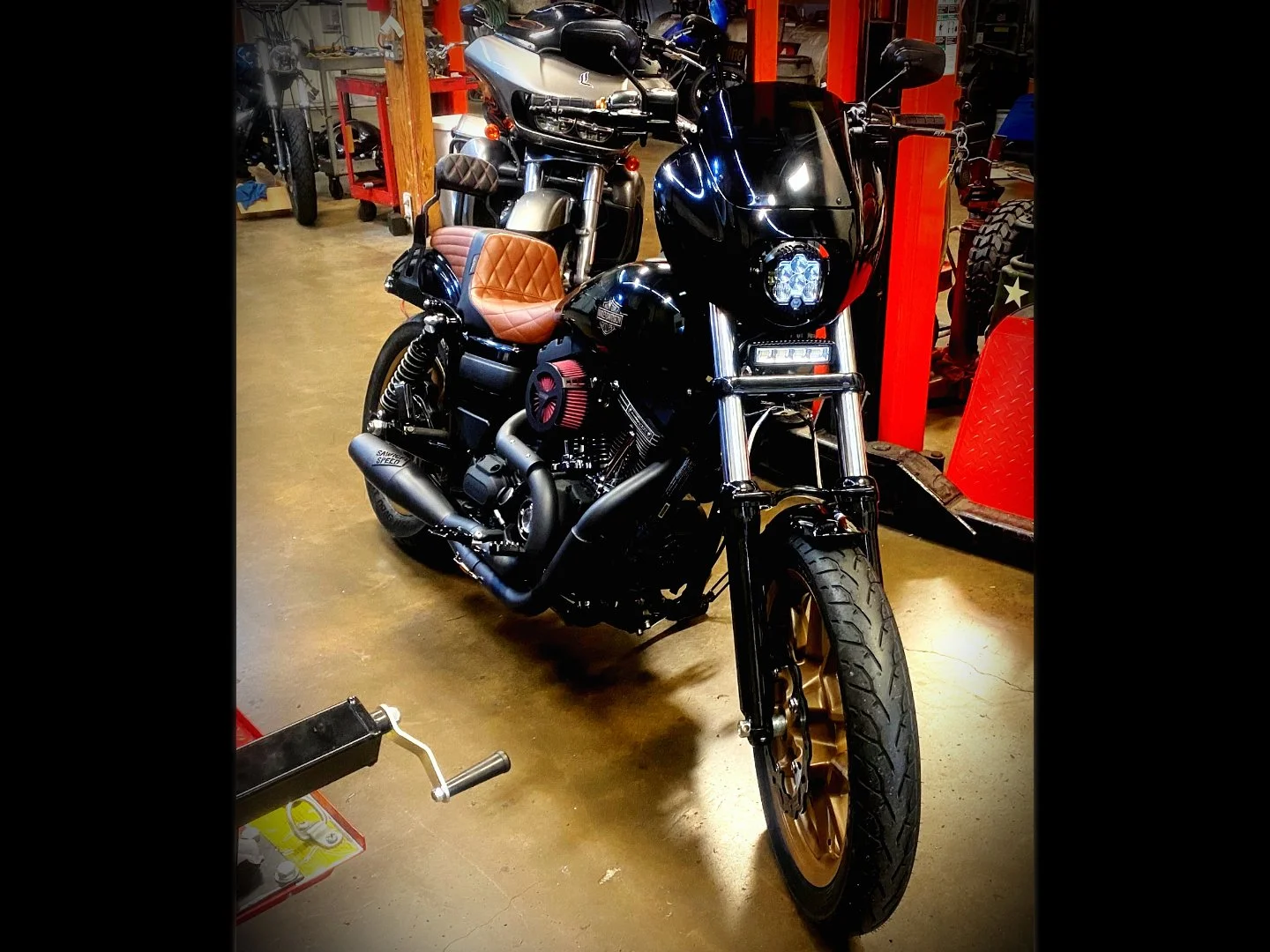 Black Harley-Davidson motorcycle with a tan quilted seat parked in a garage, surrounded by other motorcycles and tools.