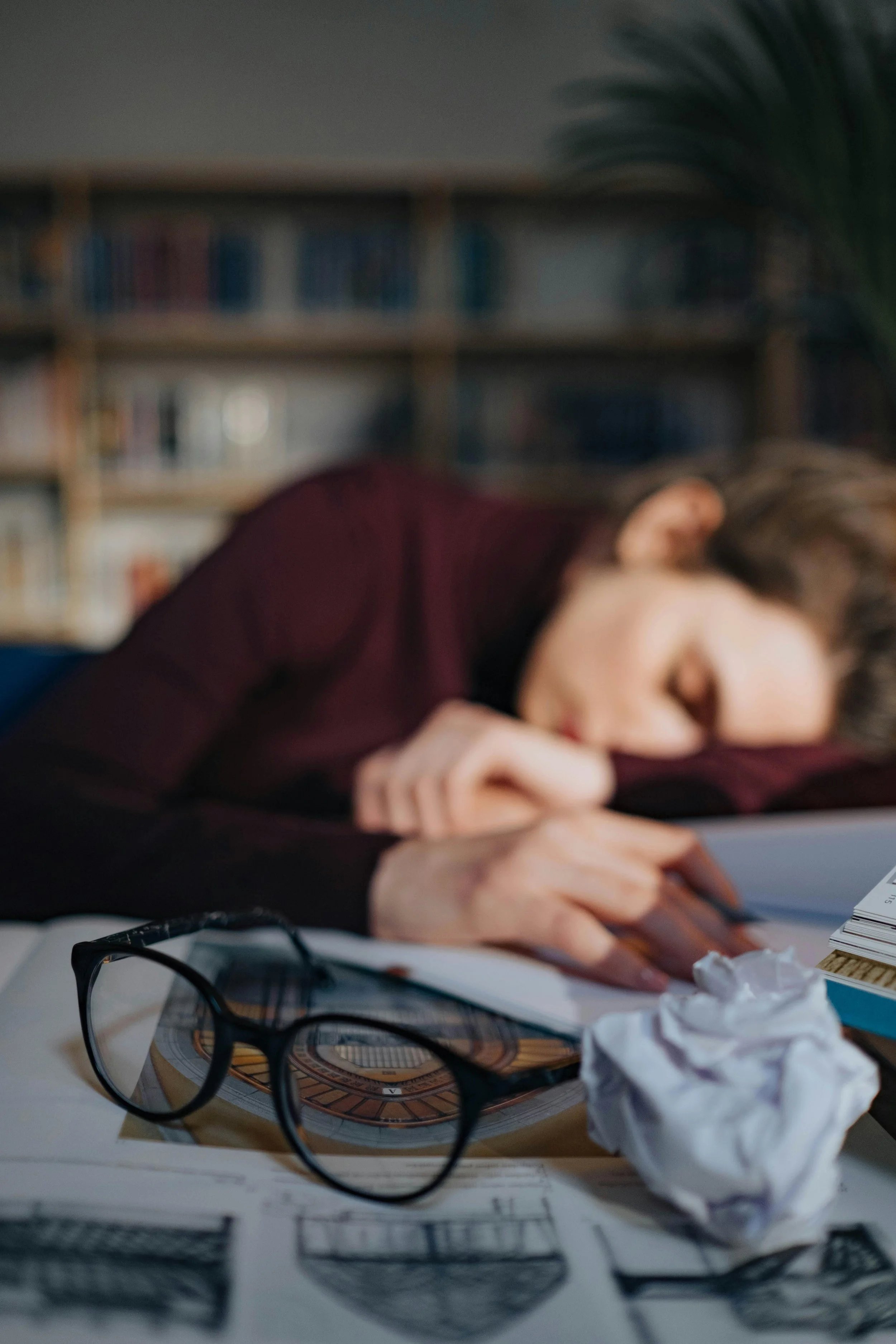A woman laying down on a messy disorganized desk, sleeping with her glasses on the table, exhausted due to ADHD burnout. Adult adhd in women can lead to severe burnout and she would benefit from virtual therapy for women with adult ADHD