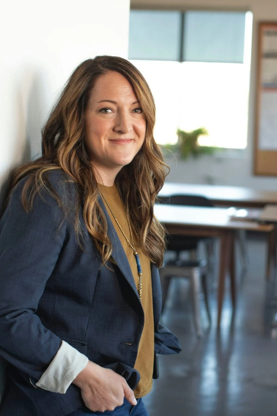 A woman with long, wavy brown hair, wearing a navy blazer, tan shirt, and layered necklace, stands inside a room with tables and chairs, smiling at the camera.