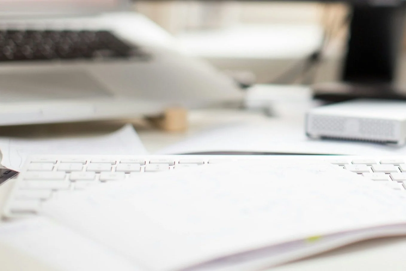 Close-up of a white computer keyboard on a cluttered desk with blurred background including a laptop and other electronic devices.