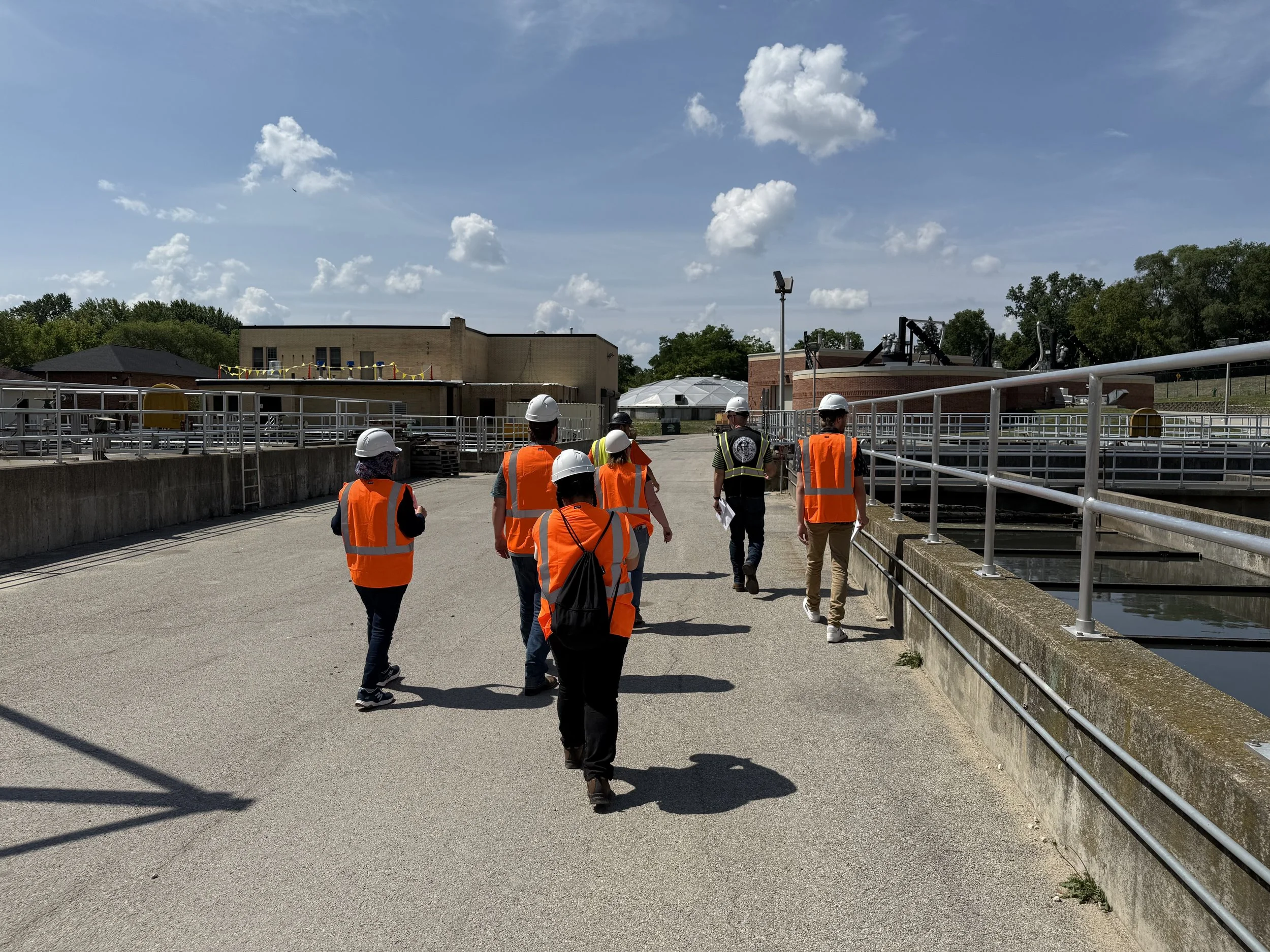 Group of people wearing safety vests and helmets walking through an industrial or water treatment facility with buildings, pipes, and water tanks.