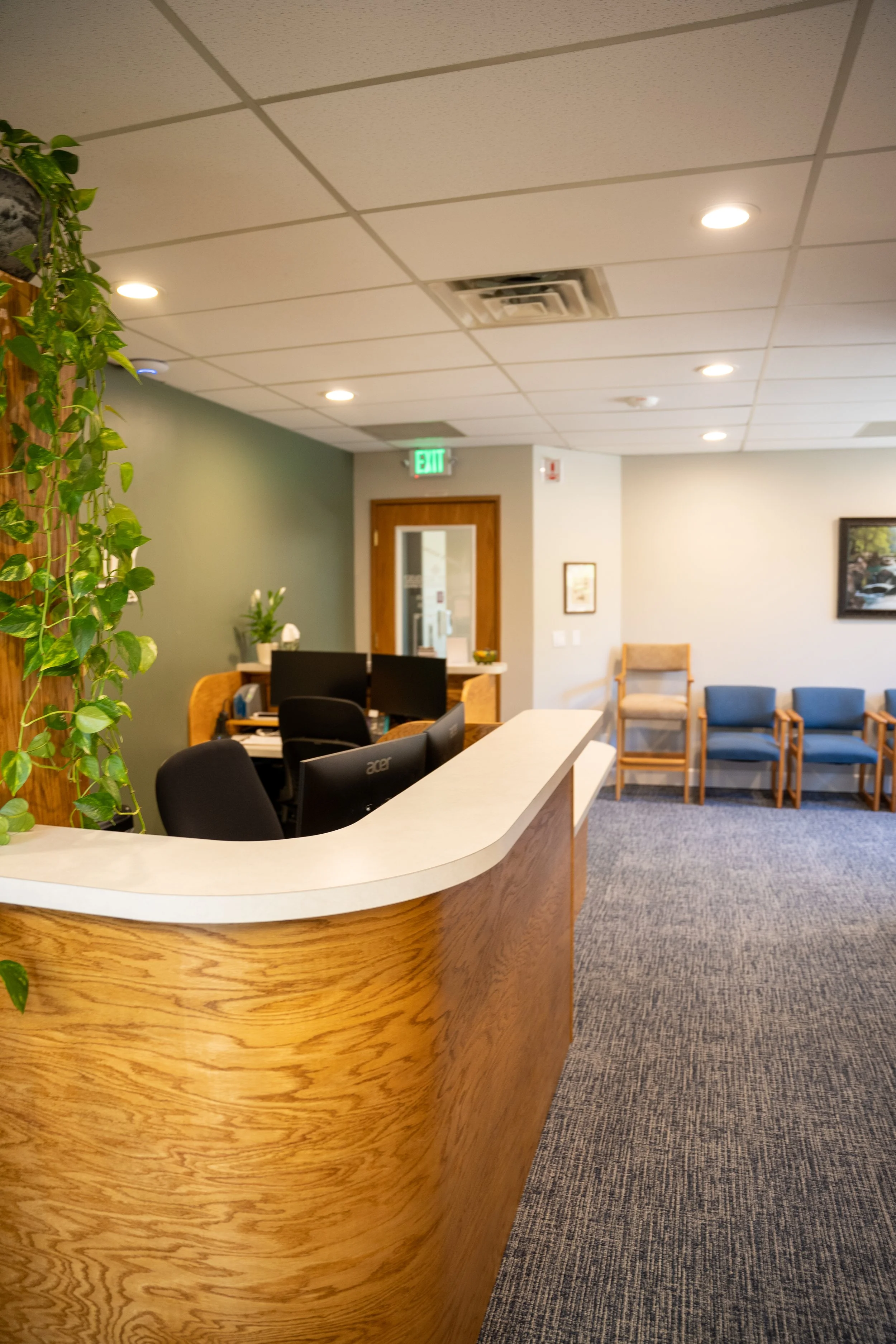 Reception area with a wooden front desk, multiple computers, green plant on the left, beige and gray chairs in the background, framed pictures on the wall, exit sign, and ceiling lights.