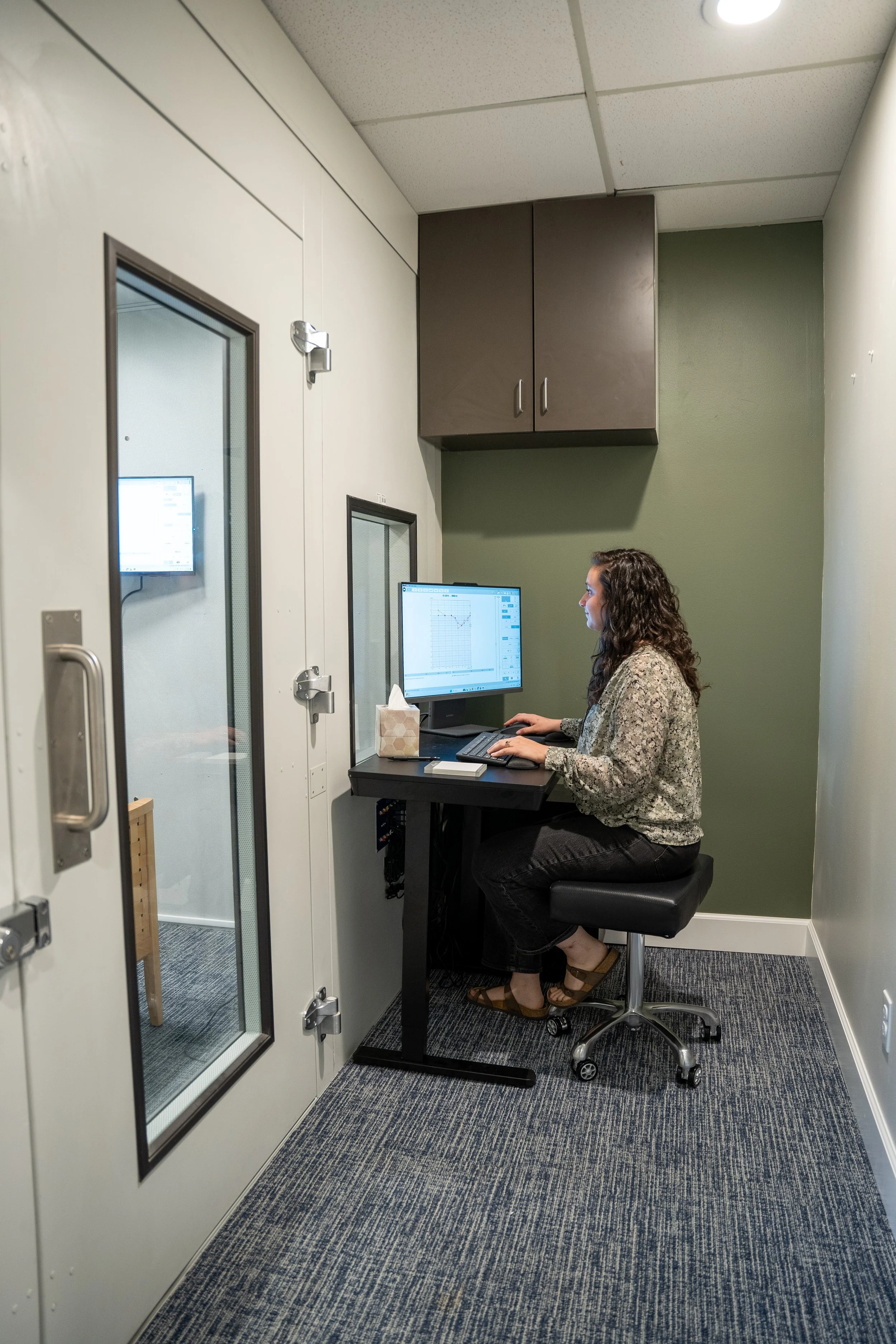 A woman sitting on a stool in a small soundproof booth, testing someone's hearing on a computer with a graph displayed on the monitor, inside an office or studio with white walls, gray carpet, and a dark brown cabinet on the wall.