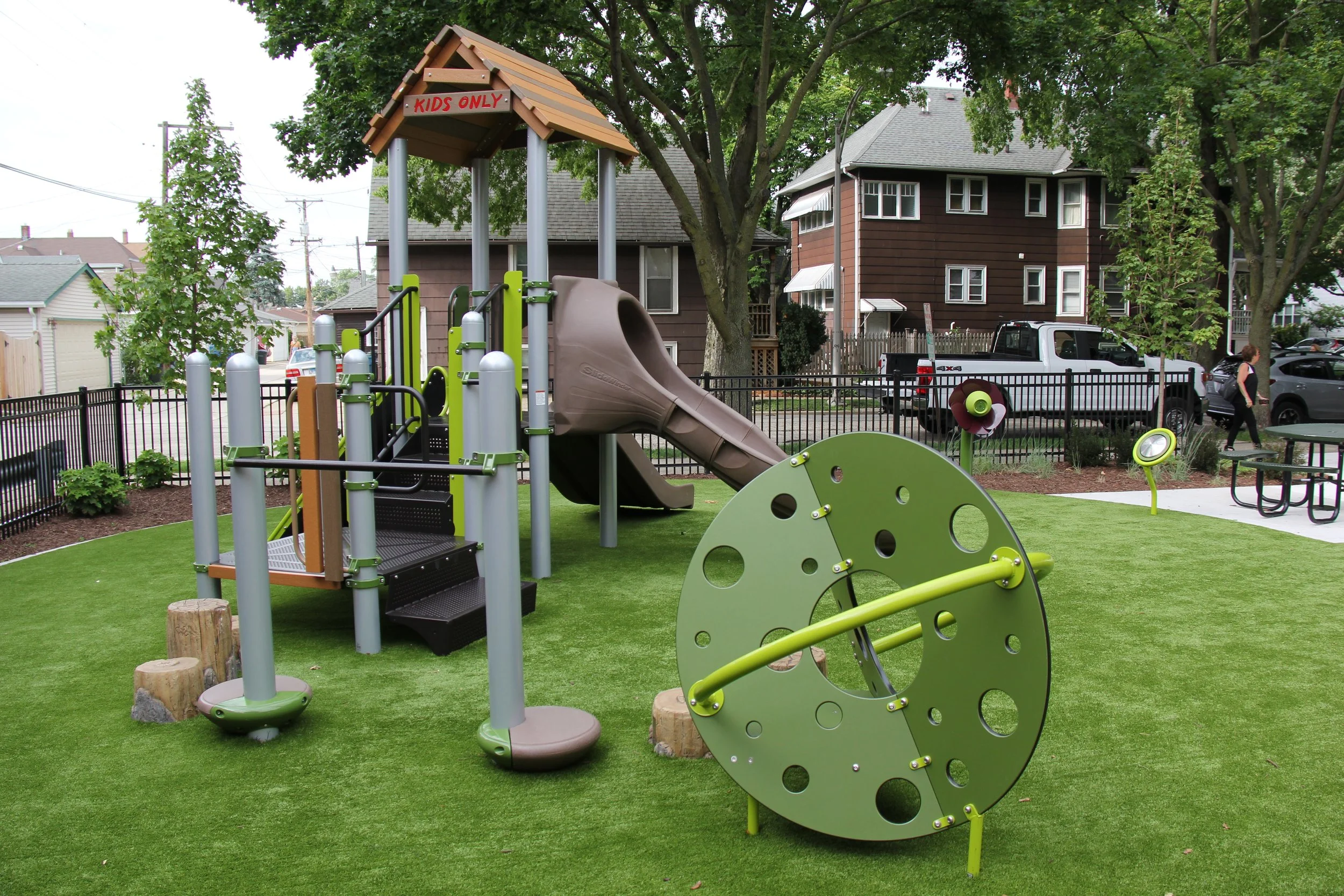 Children's playground with a slide labeled 'Kids Only,' a spinning play panel, and various playground equipment surrounded by grass and trees, with houses and parked cars in the background.