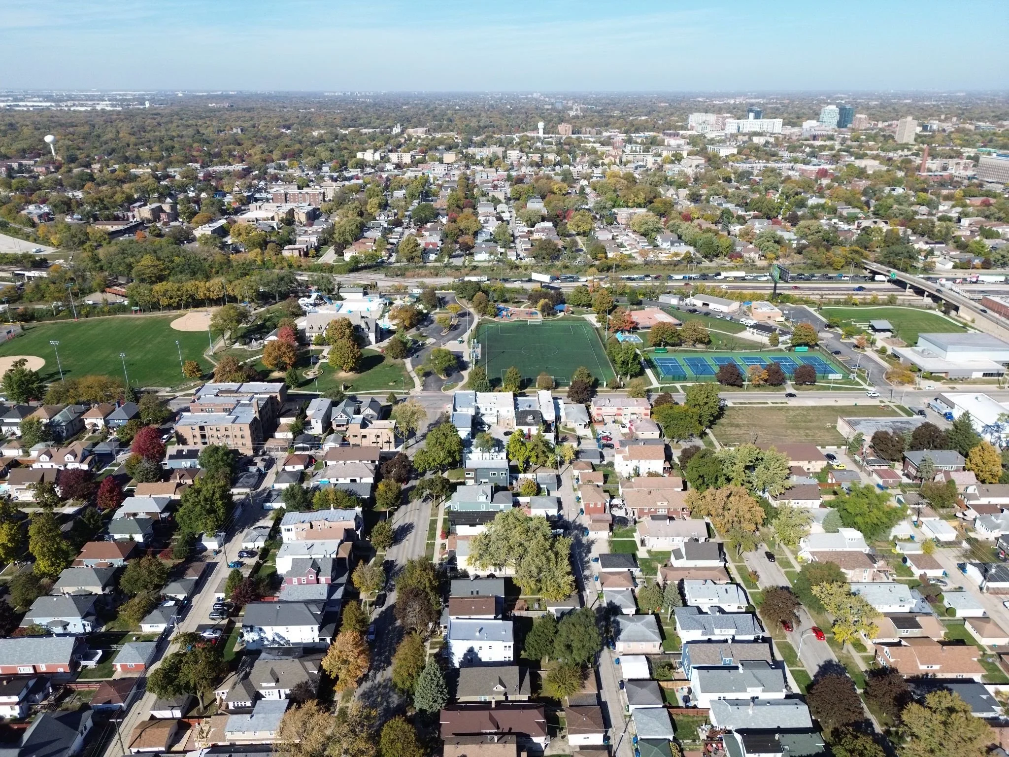 Aerial view of a residential neighborhood with houses, trees, sports fields, and a nearby highway in a cityscape.