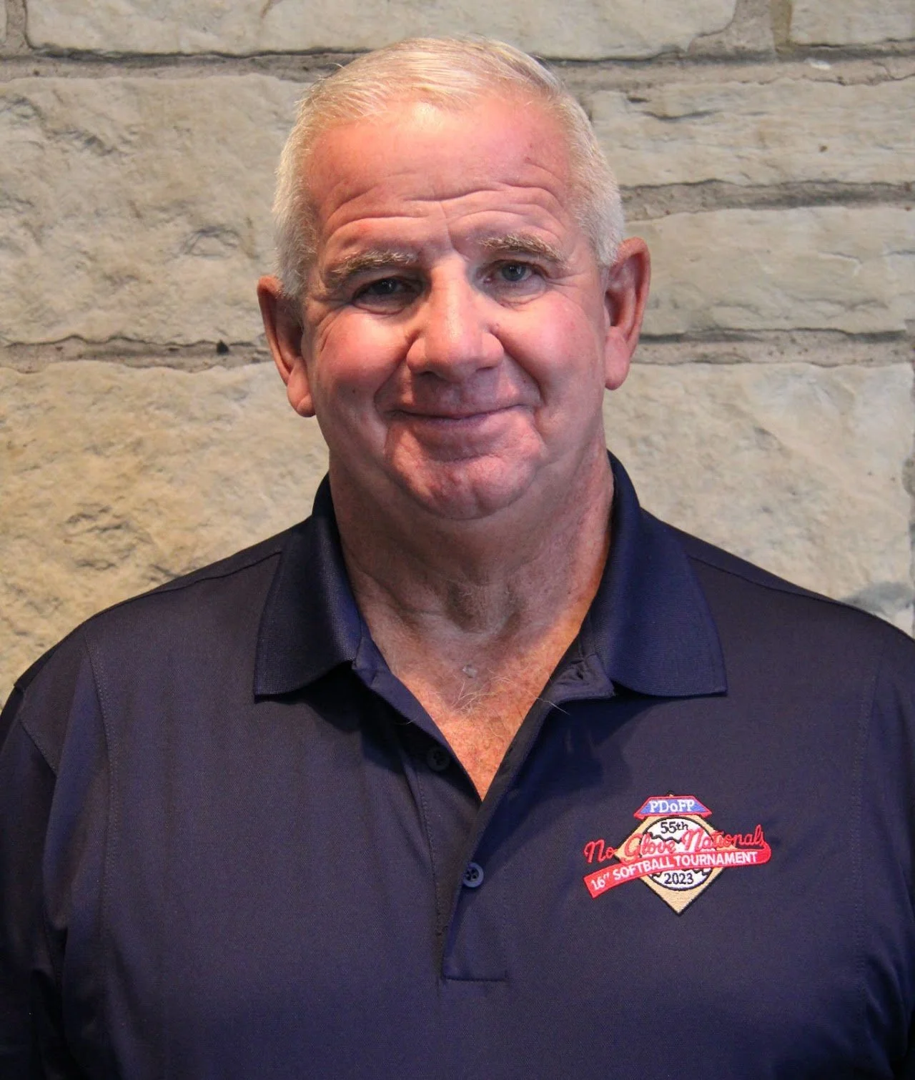 A smiling middle-aged man with gray hair wearing a navy blue polo shirt stands in front of a stone wall. The shirt has a badge with text indicating it is from a 55th National Softball Tournament in 2023.
