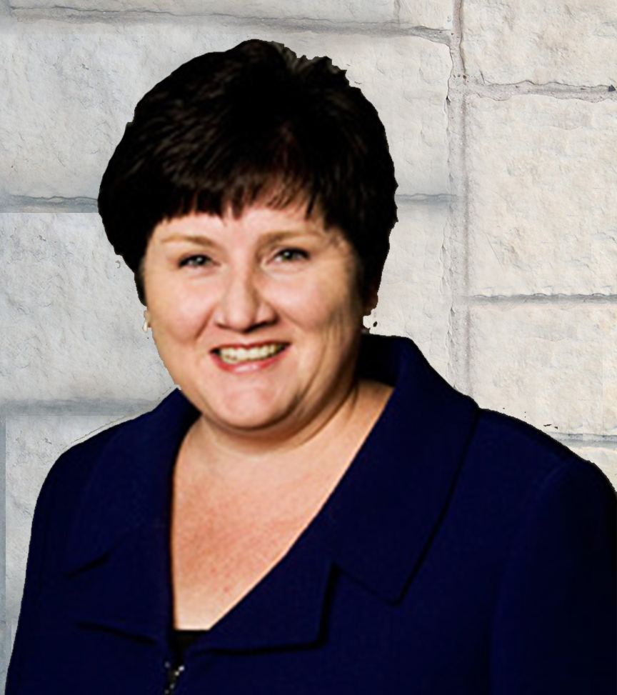 Photograph of a smiling woman with short dark hair, wearing a dark blue blazer, standing against a light-colored brick wall background.