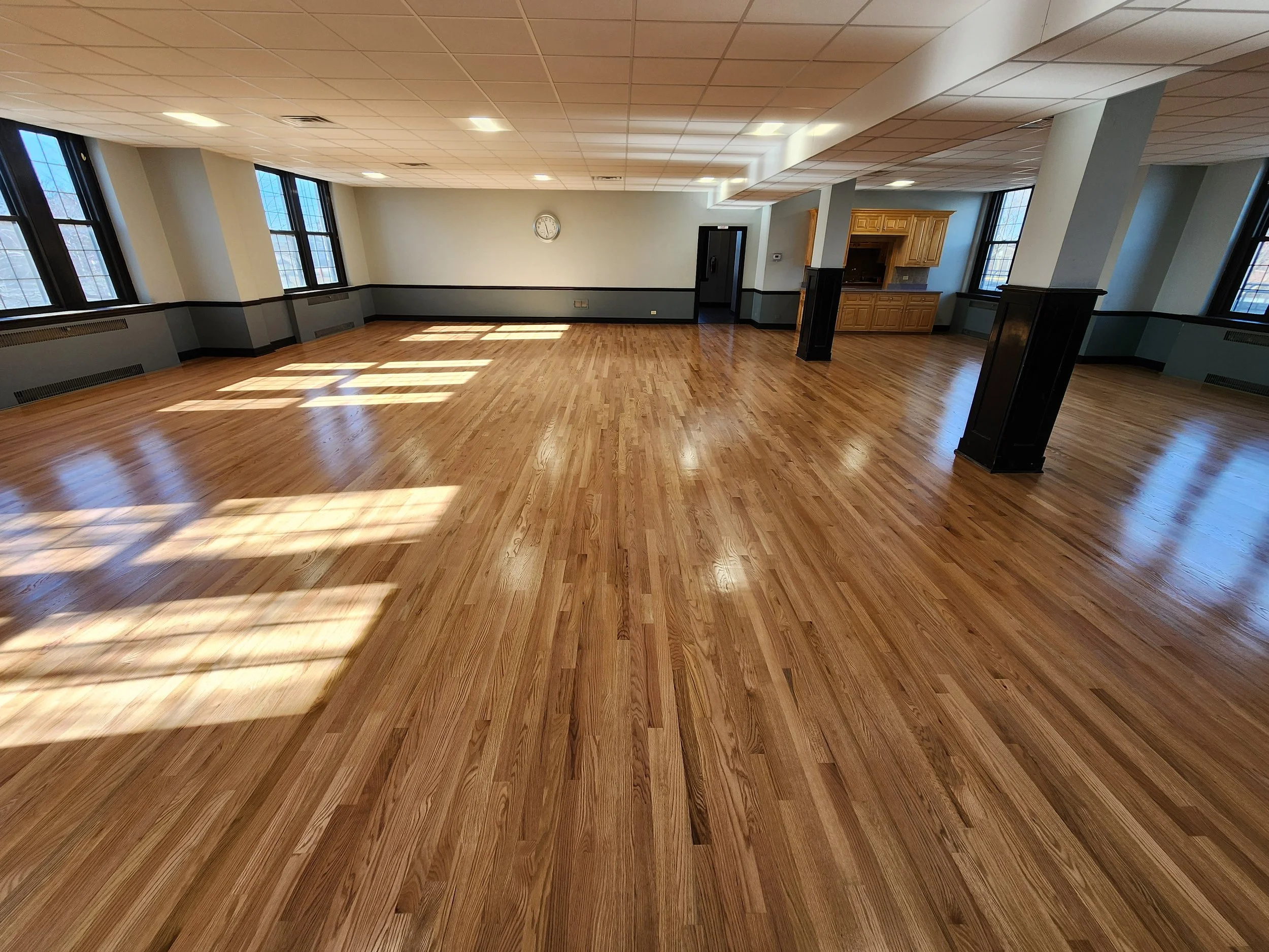 An empty room with polished hardwood floors, large windows letting in natural light, a clock on the wall, and a small kitchenette area with wooden cabinets.
