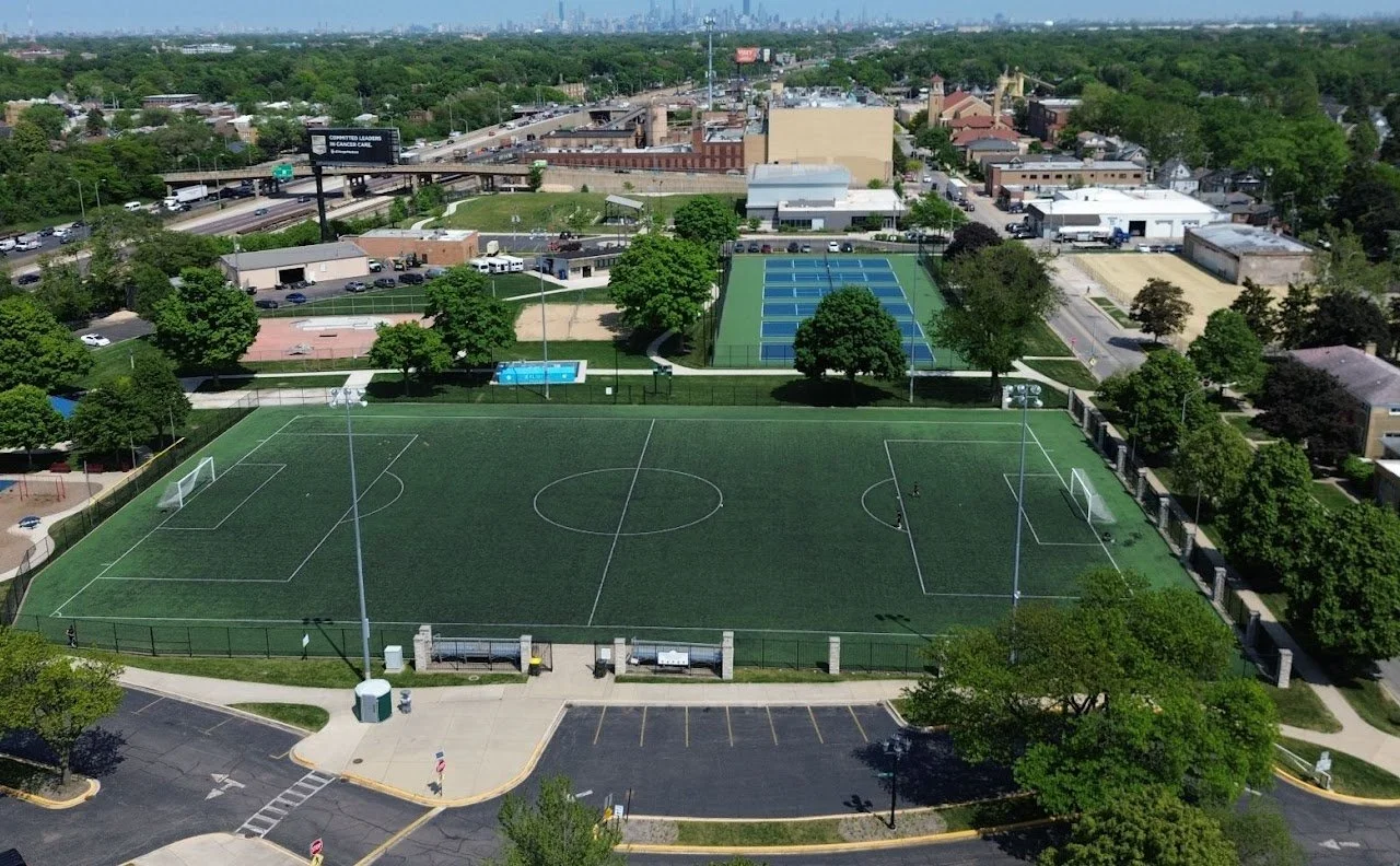 Aerial view of a sports complex including a soccer field with artificial turf, two basketball courts, and multiple tennis courts surrounded by trees and parking lots.