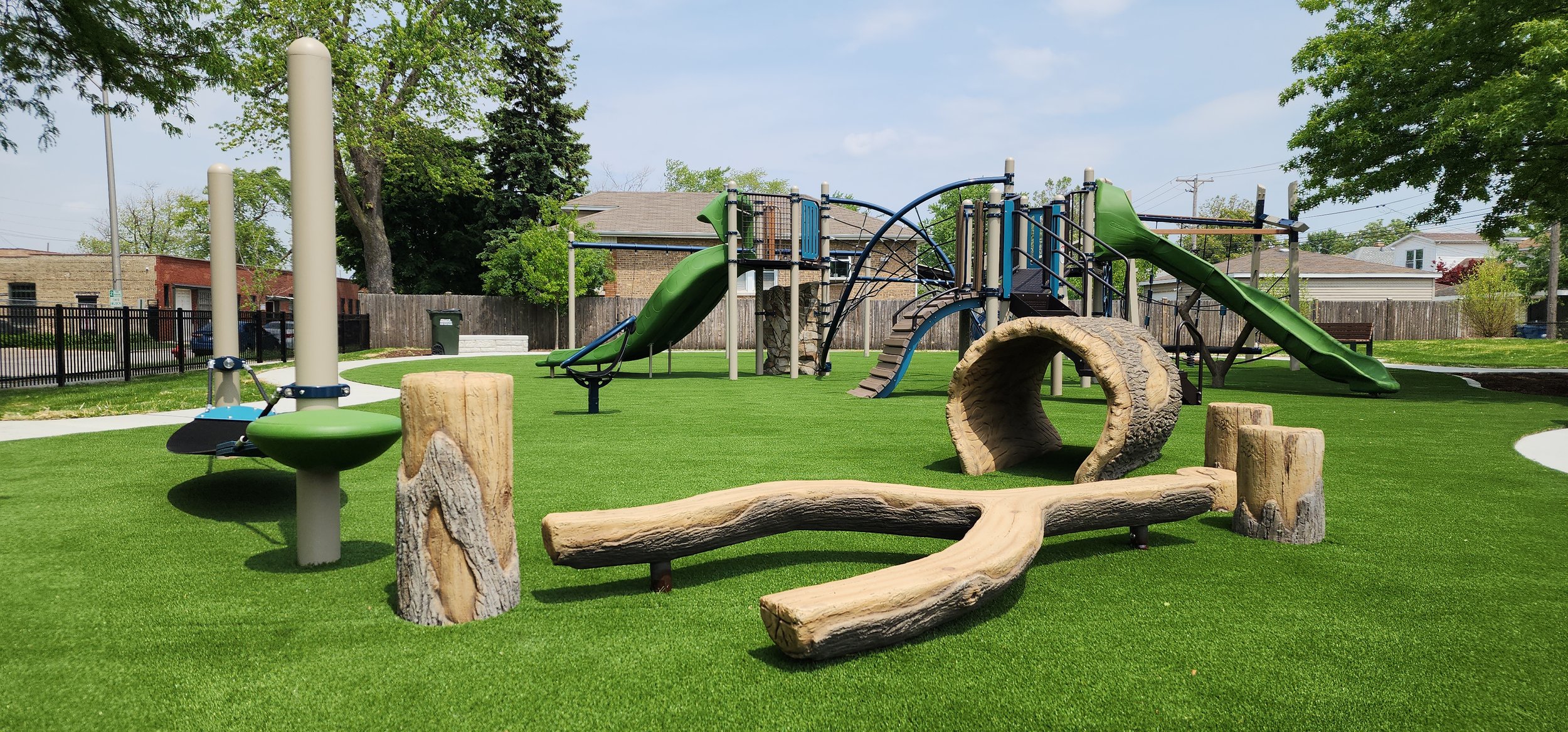 Children's playground with green grass, wood and metal play structures including slides, climbing frames, and log-shaped features in a fenced outdoor area.
