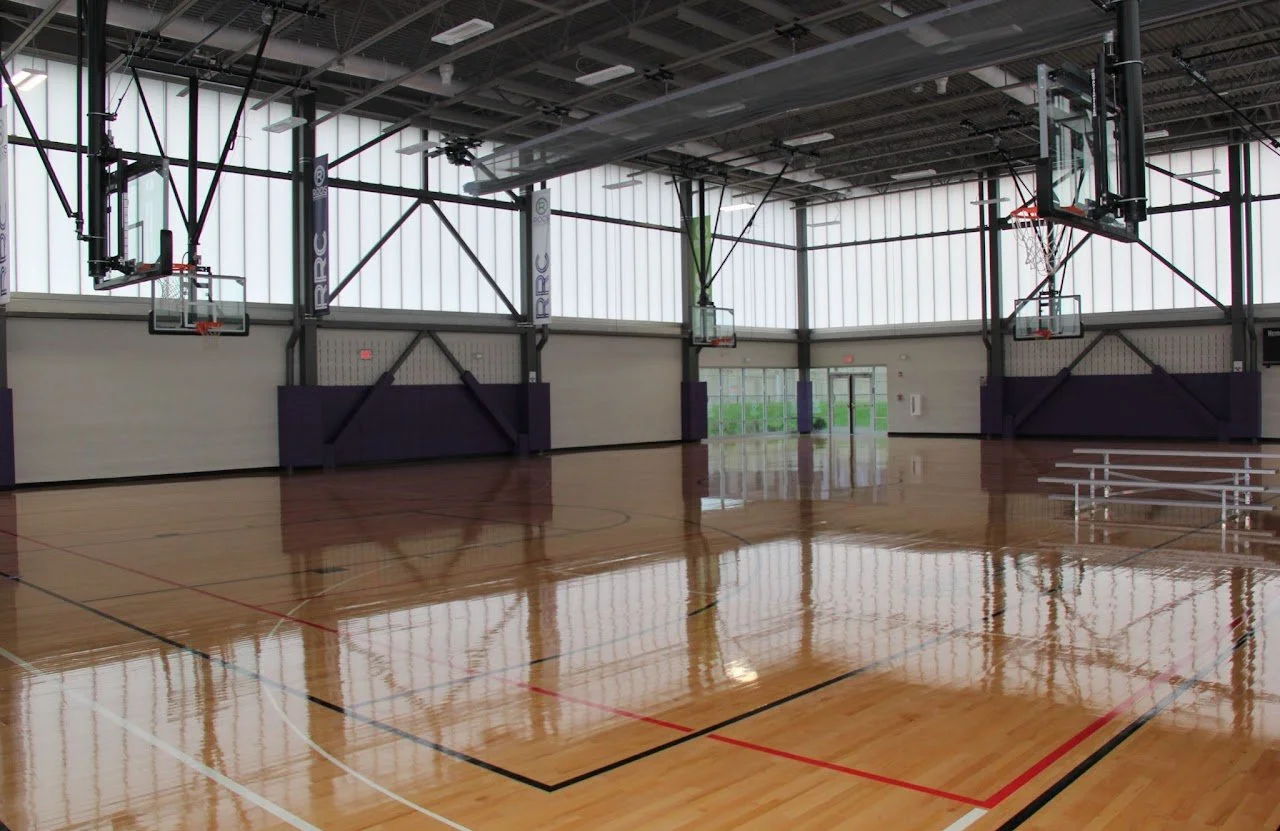 Empty indoor basketball court with polished wooden floor, three basketball hoops, and large windows allowing natural light.