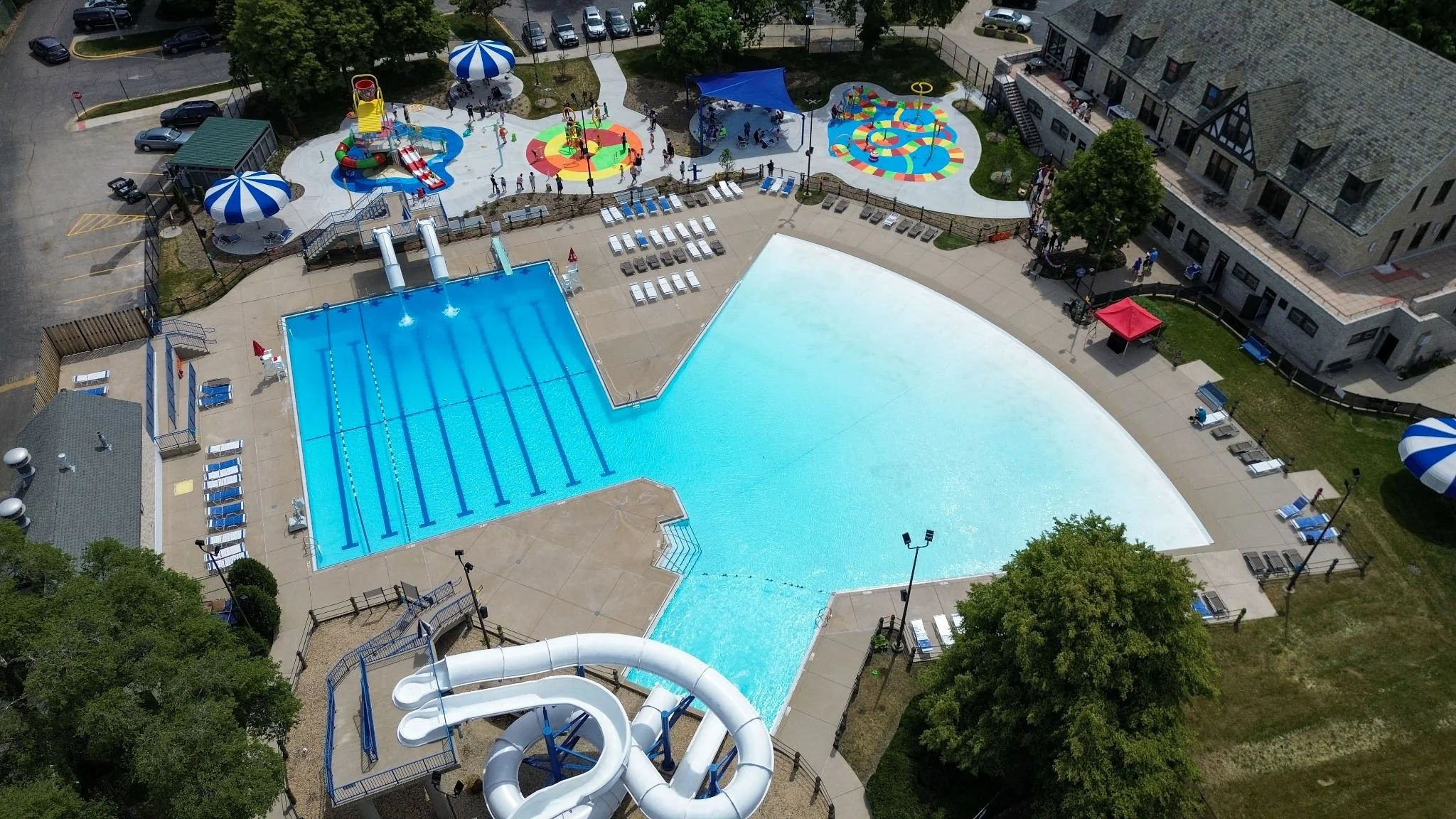 Aerial view of a colorful outdoor water park with swimming pools, water slides, a splash pad, and shaded seating areas, surrounded by trees and a building.