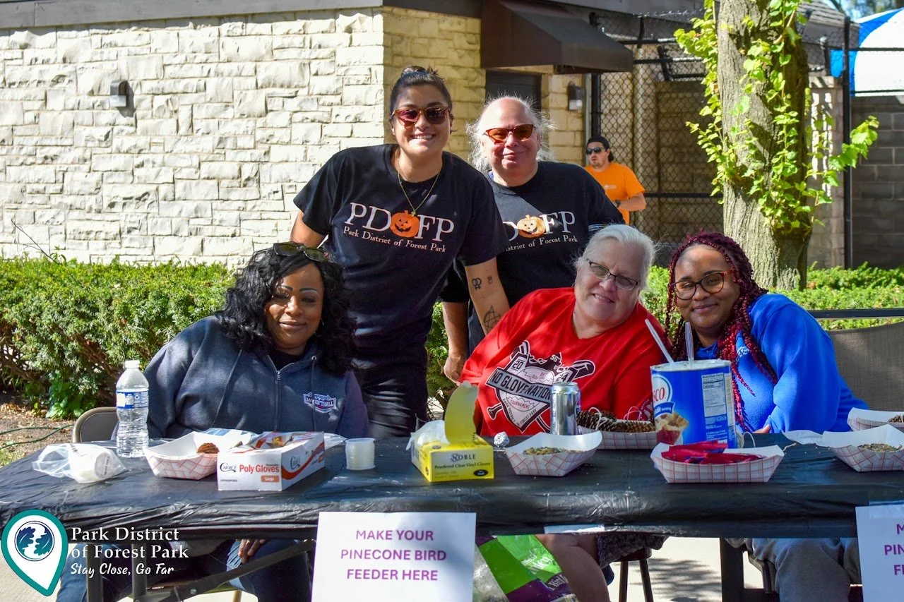 Group of five people sitting and standing at a table outdoors during a community event, with snacks and drinks on the table, in front of a white brick wall and green bushes. Sign on the table reads 'Make your pine cone bird feeder here.'