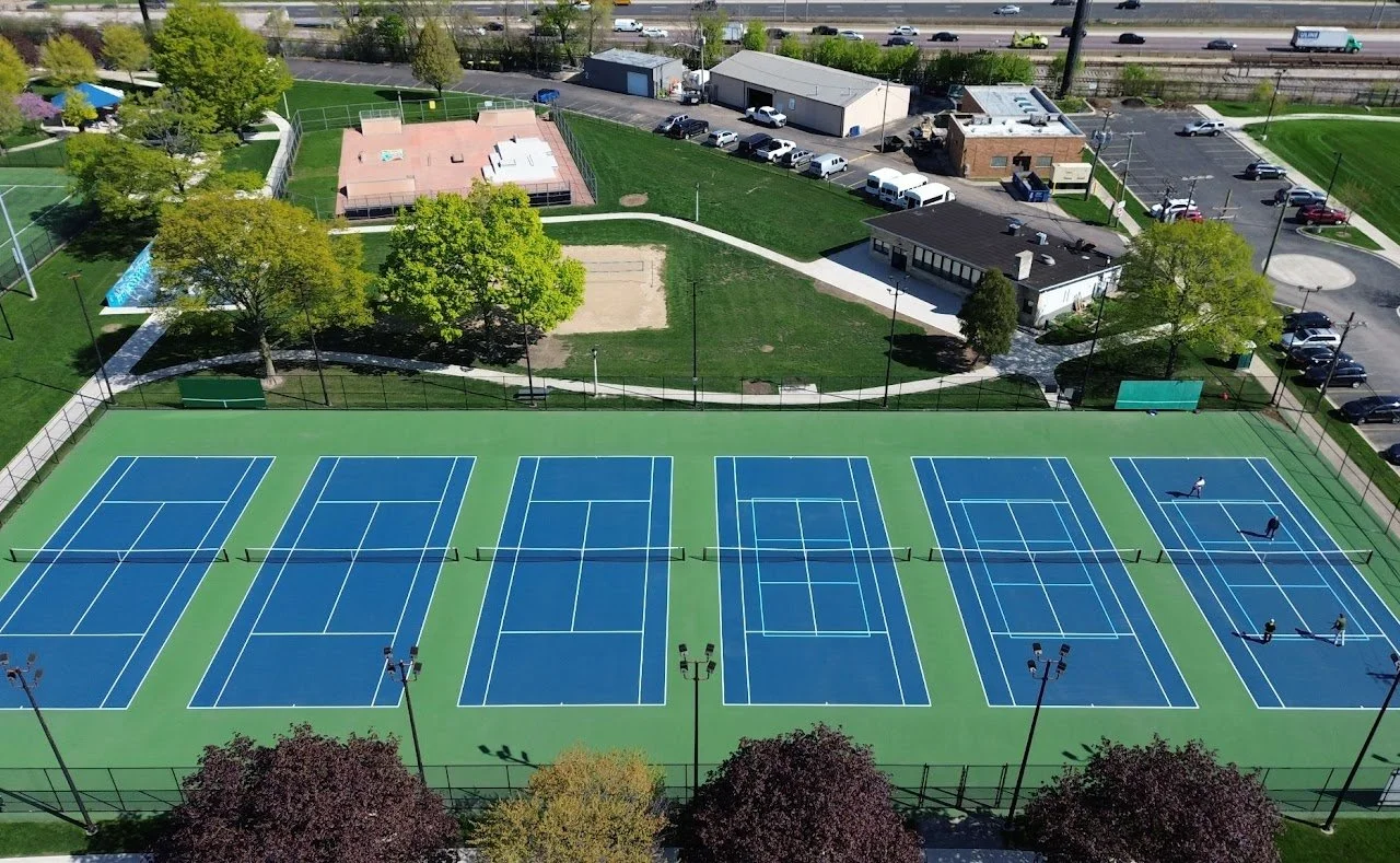 Aerial view of a sports complex featuring multiple tennis courts with blue surfaces and white lines, surrounded by a green fence. There are a few people playing tennis on one of the courts. Adjacent to the courts are trees, pathways, and buildings, with parking lot and traffic visible in the background.