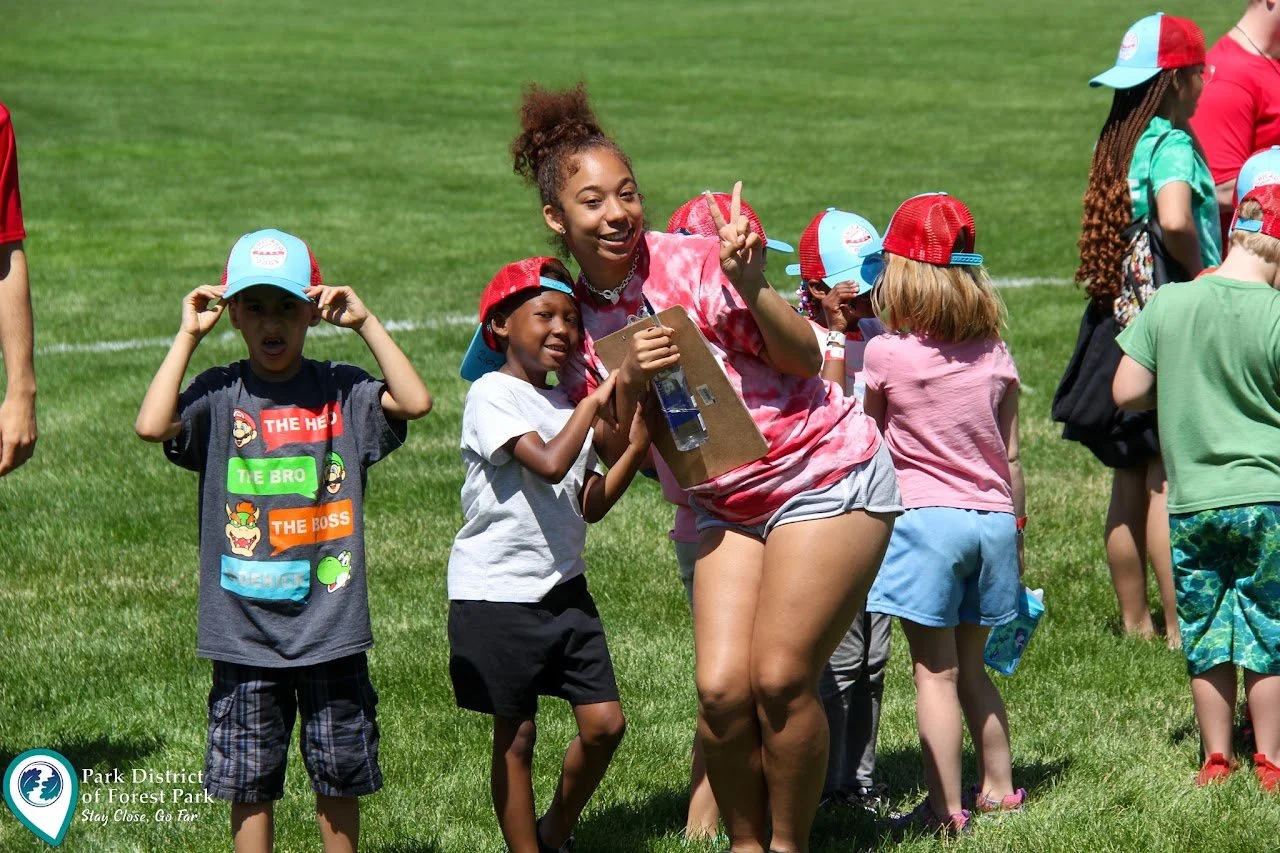A group of children and a young woman at an outdoor event on a grassy field, some wearing red and white caps, with the woman holding a clipboard and making a peace sign.