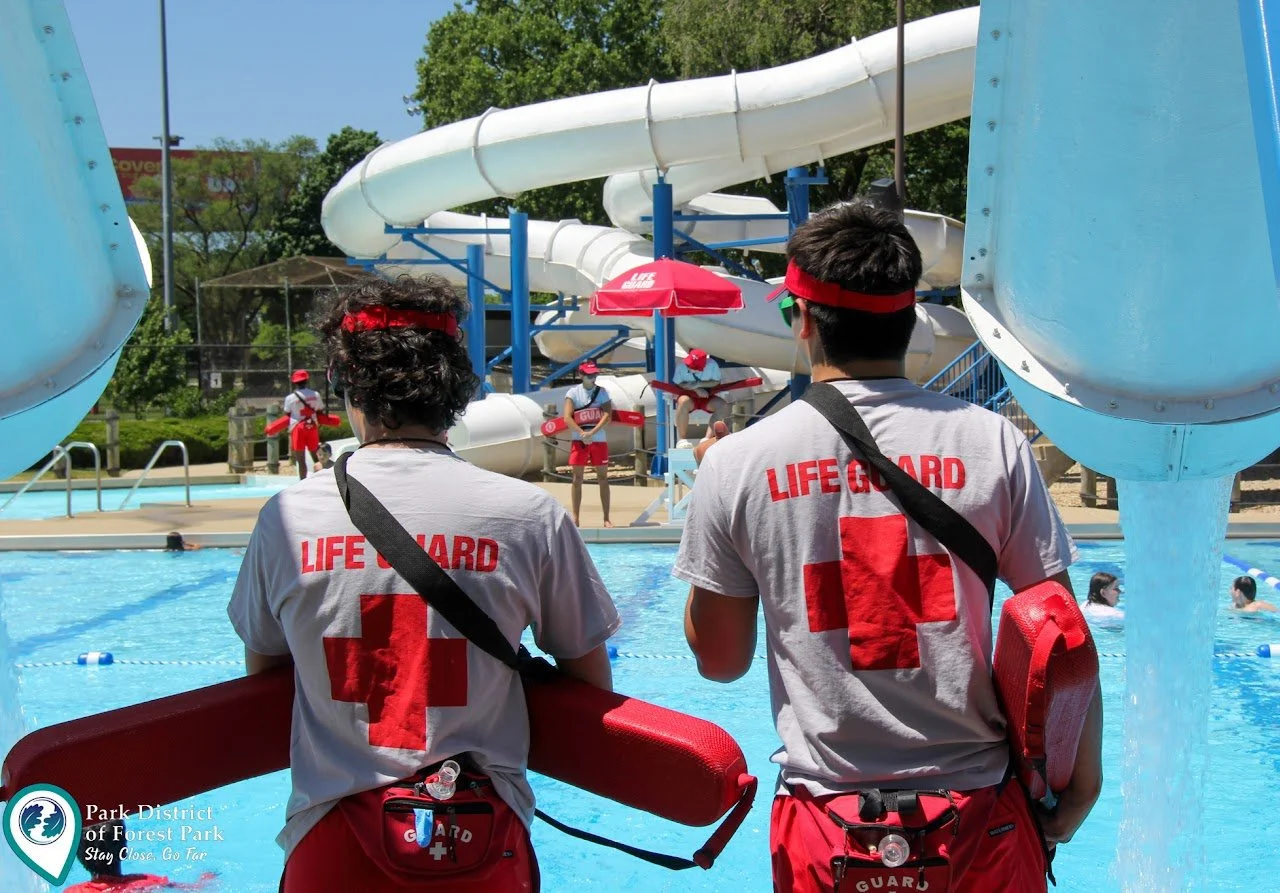 Lifeguards standing by a swimming pool, watching over swimmers, with water slides in the background.