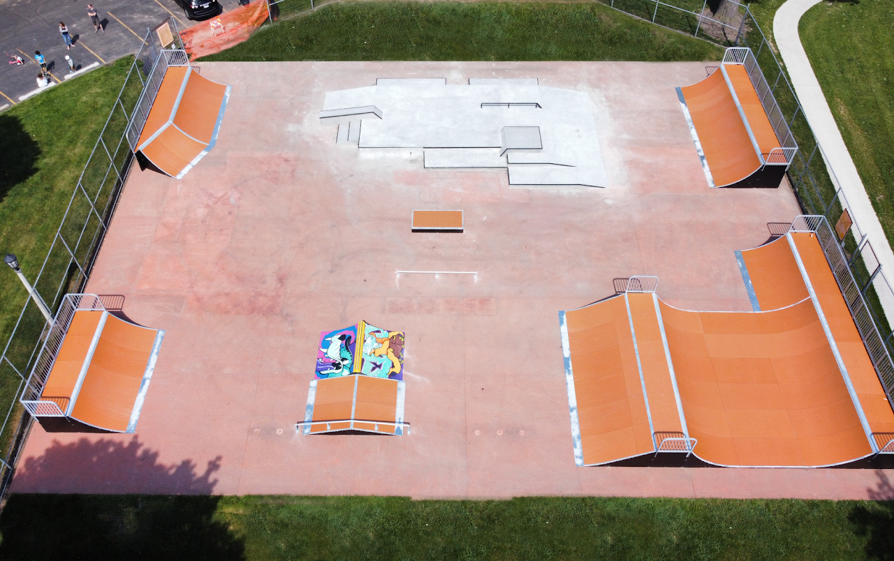 An empty outdoor skatepark with orange ramps, grind rails, and a painted area, surrounded by grass and a fence, with a few people on the adjacent parking lot.