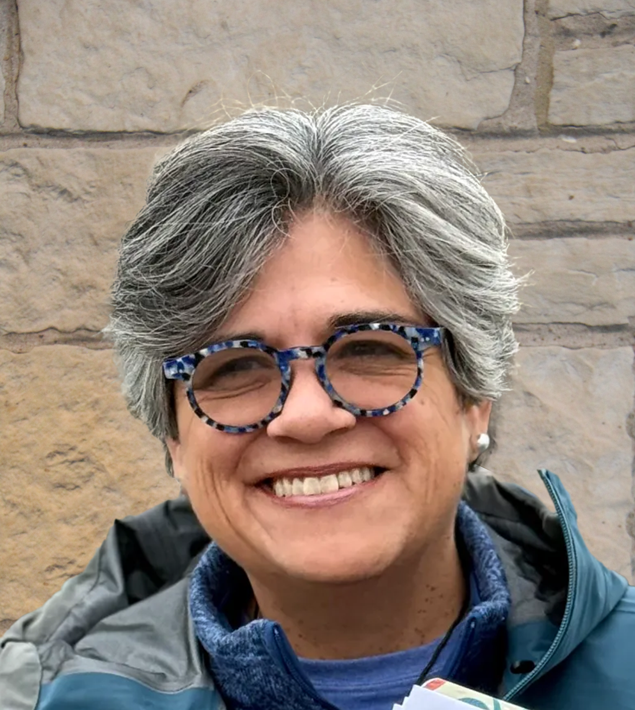 Smiling woman with short gray hair and blue patterned glasses standing against a stone wall.