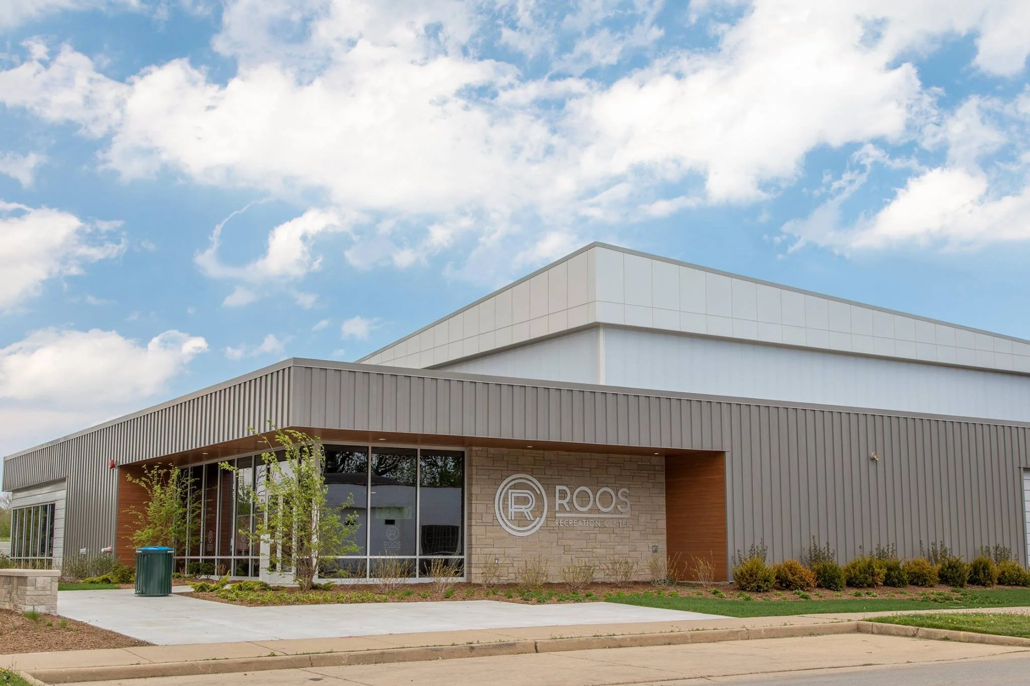 Exterior view of the Roos Recreation Center building with a clear blue sky and some clouds in the background. The building features modern architectural design with a mix of brick, wood, and metal panels, large glass windows, landscaping with small trees and bushes, and a concrete sidewalk in front.