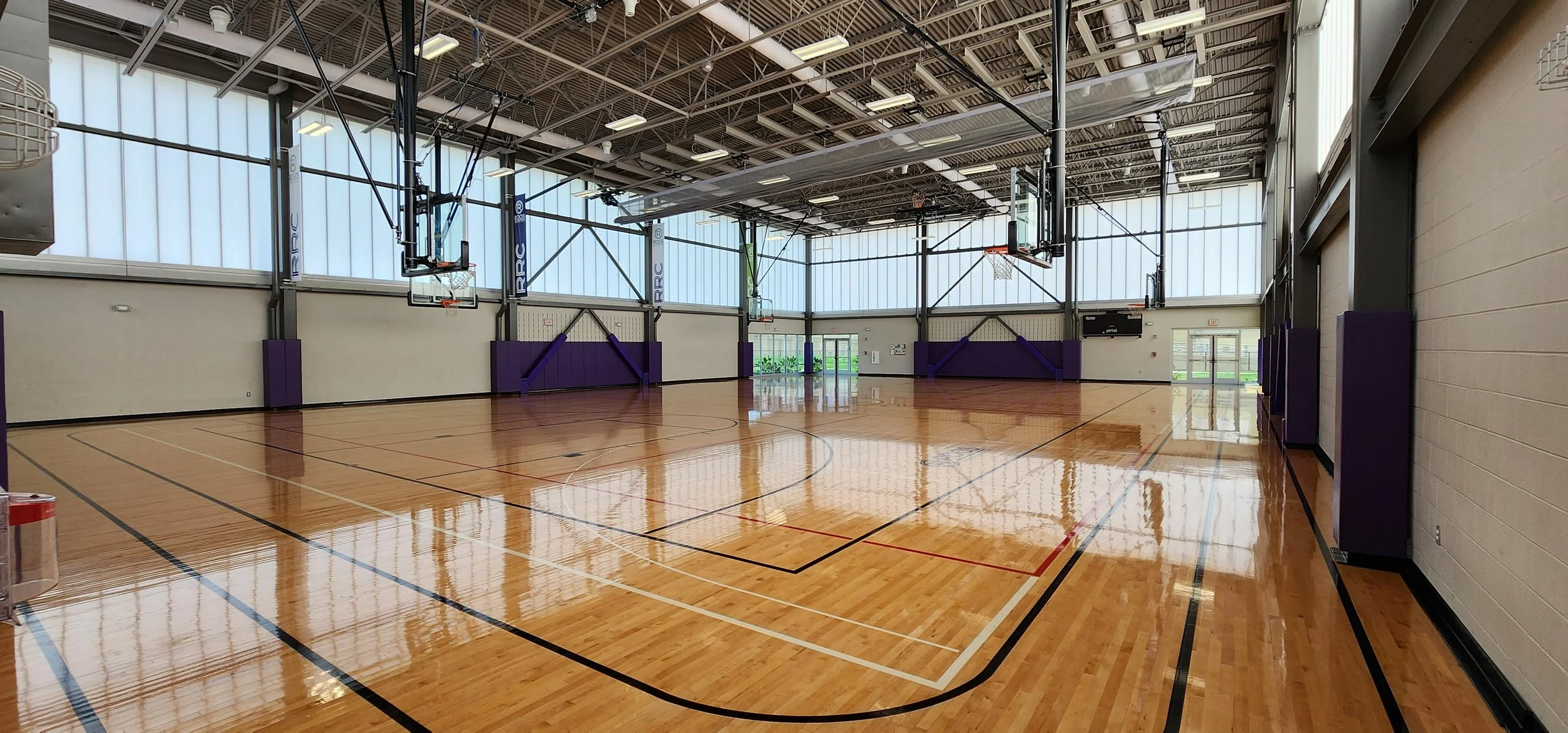 Inside a spacious basketball gym with polished wooden floor, multiple hoops, and large windows letting in natural light.