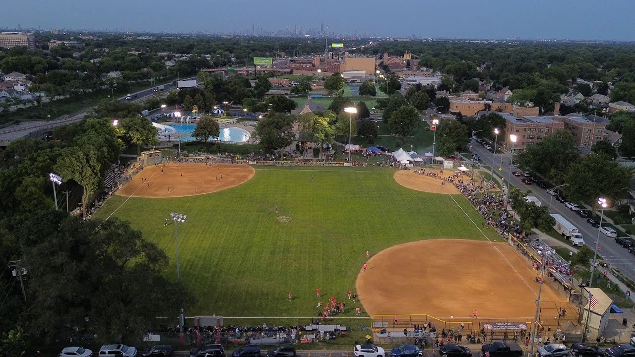 Aerial view of a baseball field with players and spectators during a game, surrounded by trees and city buildings in the background.