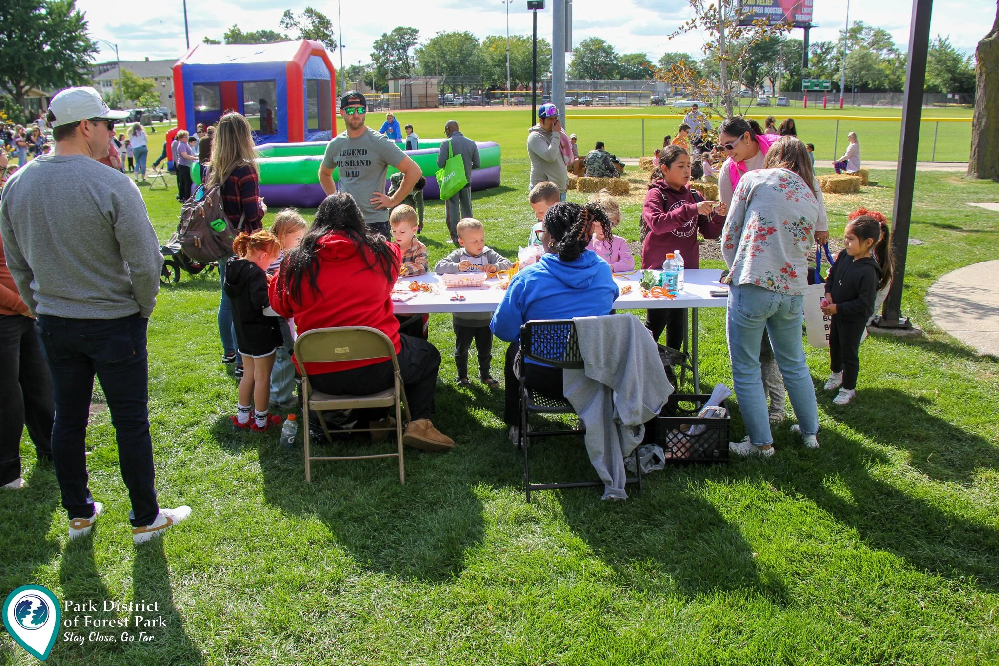 People gathered at a park for an outdoor event with children and adults sitting around a table, some standing, and a bounce house in the background.