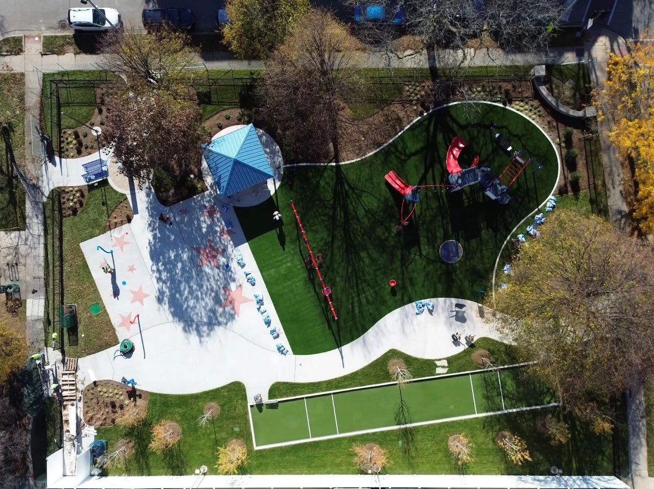 An aerial view of a playground with various play structures, a small basketball court, and landscaped areas with trees and benches.