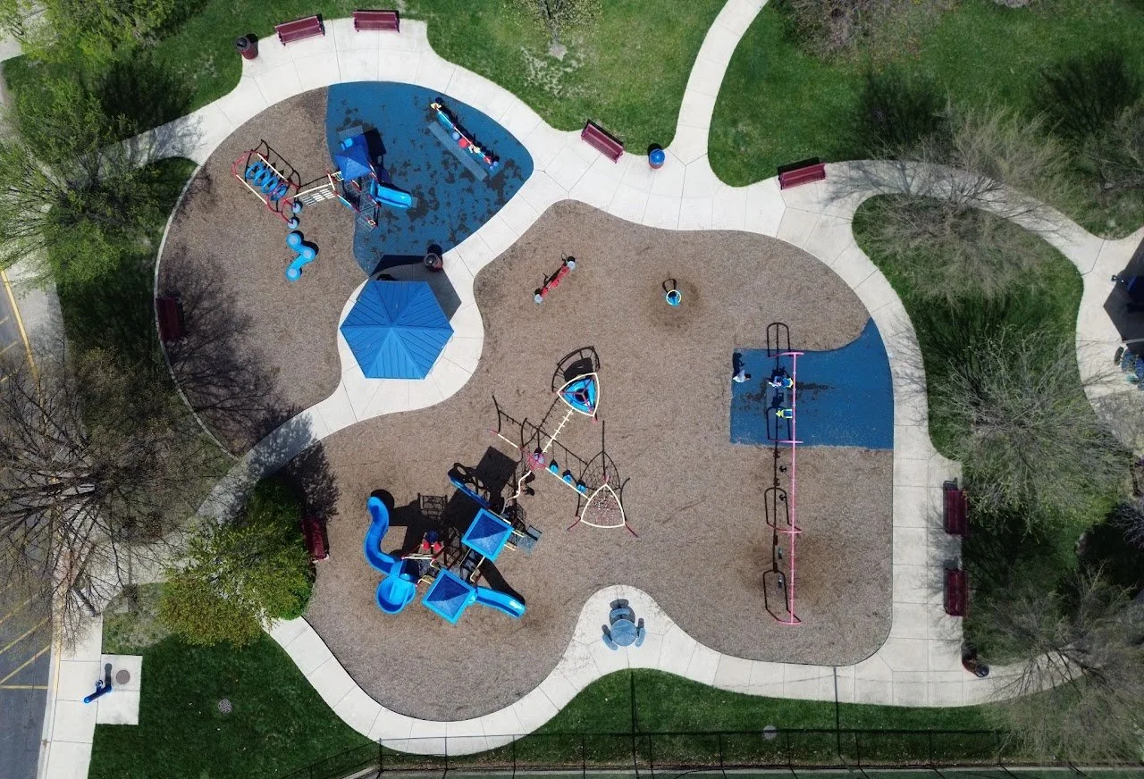 Overhead view of a playground with various play structures, slides, swings, and benches, surrounded by sidewalks, trees, and grassy areas.
