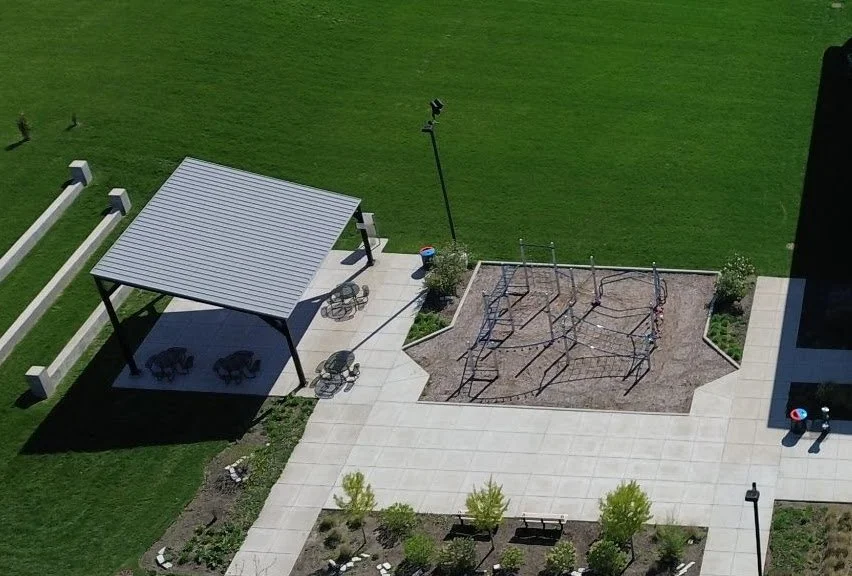 An aerial view of a backyard showing a covered seating area with chairs, a playground with climbing structures, a grassy lawn, and a paved patio with benches and trees.