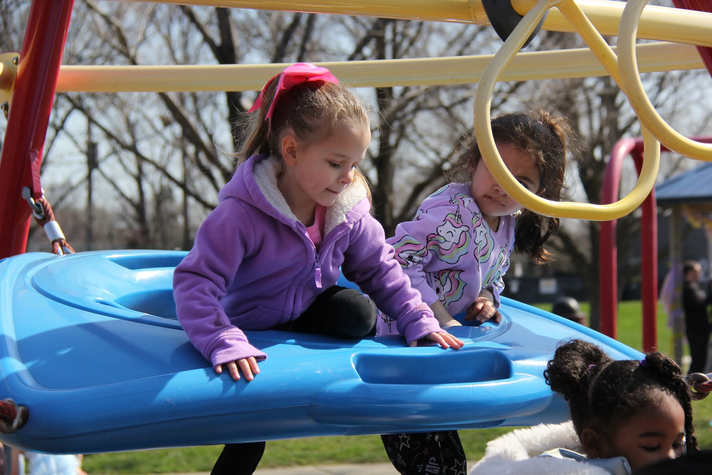 Children playing on a blue outdoor playground swing, climbing in a park with trees in the background.