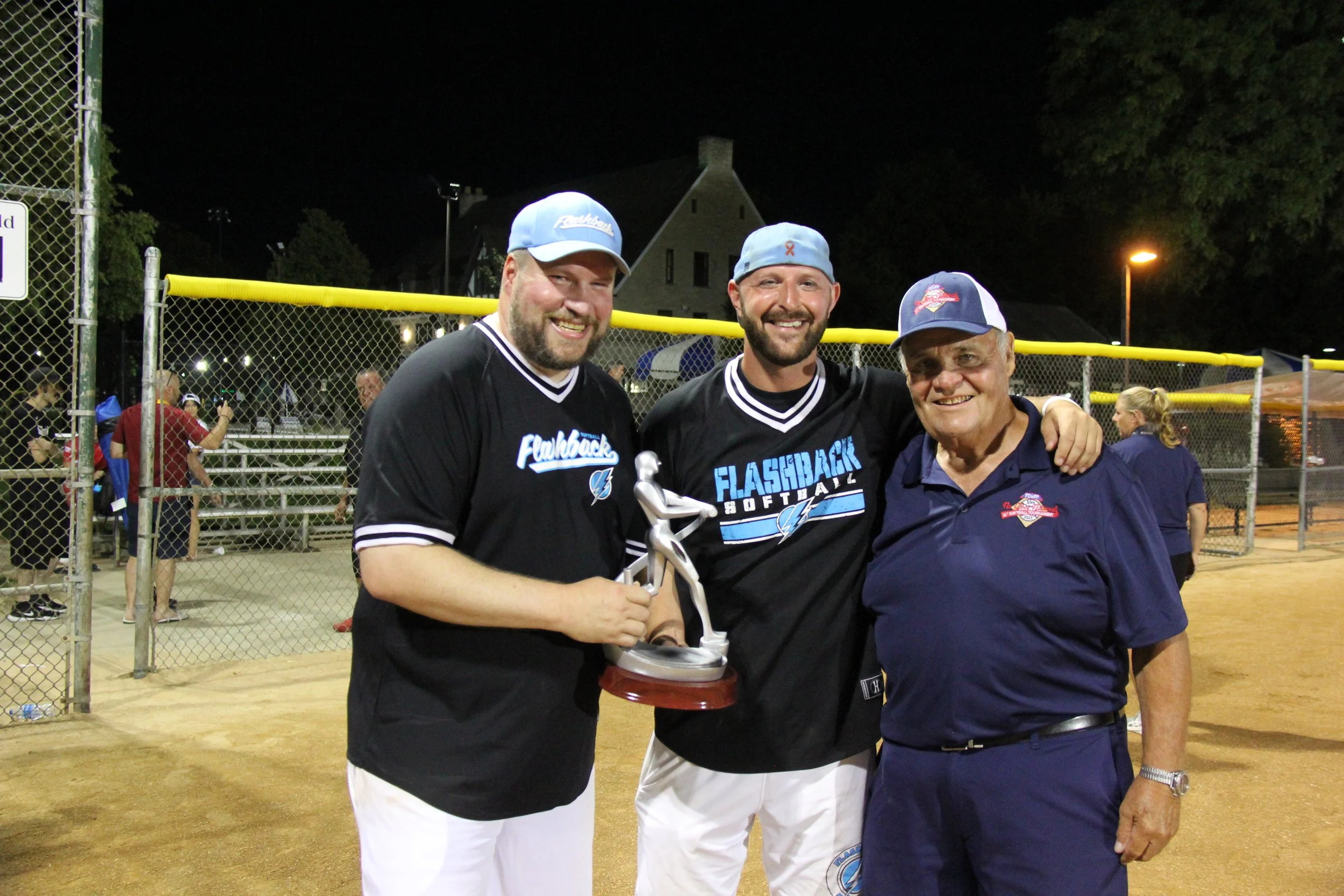 Three men smiling at night on a baseball field, with two of them wearing softball uniforms and one in a navy polo, holding a trophy, with a chain-linked fence and bleachers in the background.