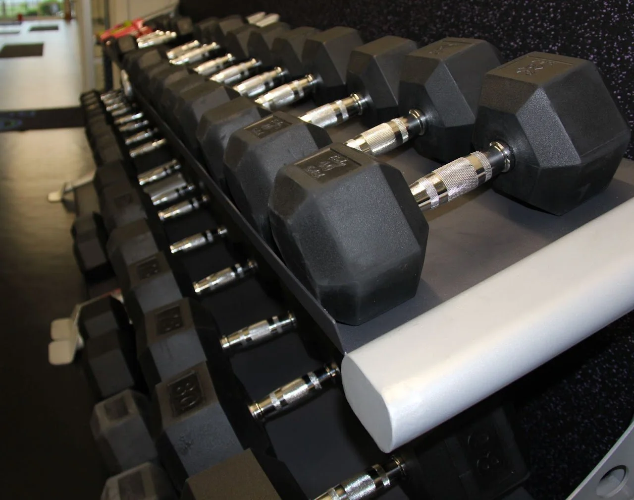 A row of black dumbbells with chrome handles stored on a black rack in a fitness gym.