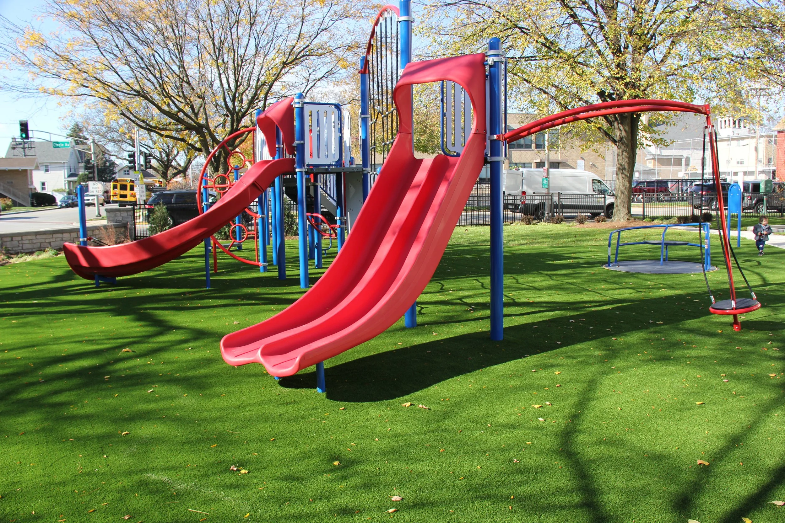 Colorful playground with red slides, a blue climbing structure, and swings, set on green grass in a park with trees and a fence in the background.